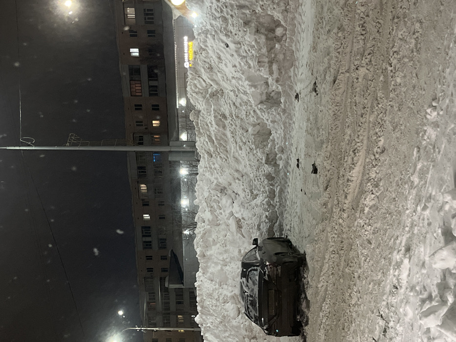 The image depicts a nocturnal, snow-covered urban scene in Neftekamsk, Russia. A dark-colored Mercedes-Benz sedan is positioned at an angle in a snow-laden area, its rear facing the camera. The vehicle's left rear is situated against a substantial, irregular snowdrift that extends across the midground. The sedan's rear bumper area exhibits significant discoloration or impact damage. Scattered dark, indistinct debris is visible on the packed snow directly in front of the large snowdrift and near the vehicle. In the background, a multi-story residential building with several illuminated windows is visible above a lower commercial structure. This commercial building features a sign with Cyrillic text, including "СОВА БЕЛЫЙ" (White Owl) and "ПРОДУКТЫ" (Products), and is brightly lit. Multiple streetlights and a tall utility pole provide artificial illumination to the scene. Visible white specks in the air suggest active snowfall. No individuals are present.