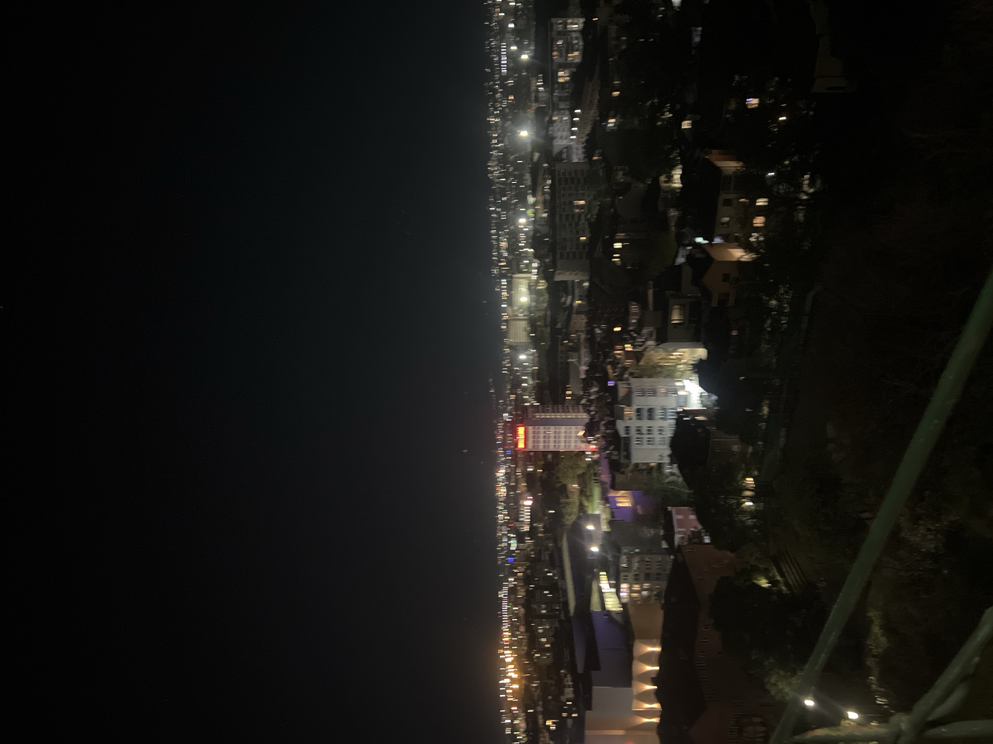 A nighttime, high-angle view of the city of Plovdiv, Bulgaria. The city stretches from left to right, illuminated by streetlights and building lights. A large, dark area occupies the left side of the image, suggesting a vast, unlit space, possibly a park or open area. In the center, a cluster of tall buildings stands out, with one building displaying a sign in red letters. The foreground is dark and indistinct, with hints of greenery and metallic structures, suggesting a viewpoint from a high location, perhaps a tower or hill.