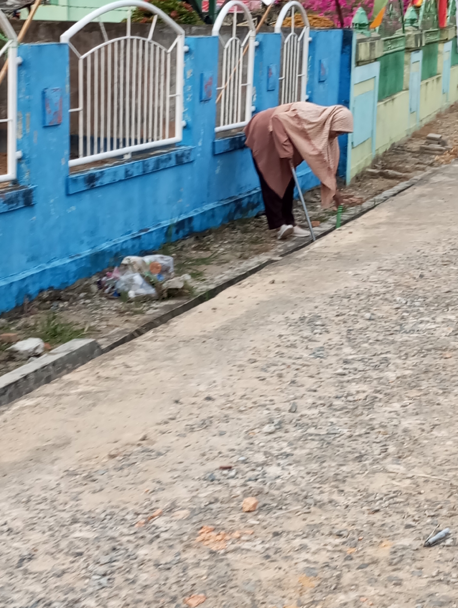 In Pematang Reba, Indonesia, a person wearing a light brown hijab and a dark garment bends down, using a stick to poke at debris near a blue wall. The wall has white metal fencing in front of it, and beyond the wall, some greenery and colorful flags are visible. To the right of the person, a section of a light yellow and green wall runs alongside the scene. The ground in the foreground is rough and stony, with a concrete edge running along the bottom left. Trash is visible on the ground near the blue wall.