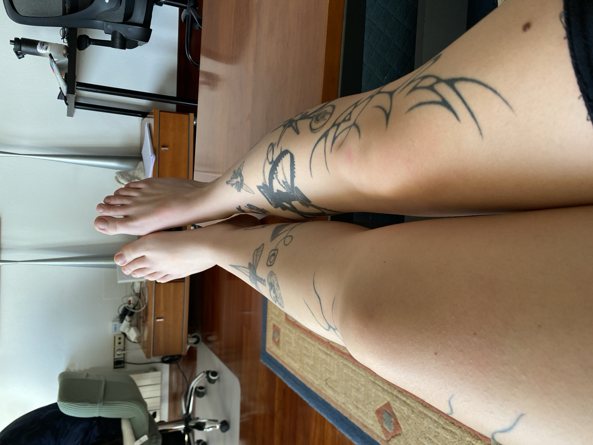 The image depicts a first-person perspective of a person's bare lower legs and feet, resting horizontally on a polished wooden surface, likely a desk or table, within an indoor environment in Gijón/Xixón, Spain. The subject's left leg (viewer's right) exhibits multiple black-ink tattoos, including a depiction of a dragonfly, a skull, and various abstract, geometric, and linear designs. The subject's right leg (viewer's left) is also extensively tattooed with black-ink designs, featuring prominent abstract and tribal-style patterns, including a large, dark, stylized motif on the calf and circular elements higher on the thigh. Both feet are visible with all toes.
The immediate foreground is dominated by the subject's legs and the wooden table surface. Below the table, a wooden floor is partially covered by a rectangular rug with a blue border and a beige central area featuring brown and red geometric patterns. In the midground, to the left, a light green upholstered office chair with chrome five-star base and wheels is visible. Behind it, against a white wall, is a light brown wooden cabinet or chest of drawers on wheels, upon which various electronic cables, adapters, and a power strip are cluttered. A tall, slender, silver-colored floor lamp stands between the cabinet and another darker wooden table. To the right of the subject's legs, a black office chair with a mesh back is visible, positioned behind a black table or desk holding a black water bottle, a small white notebook, and a pair of eyeglasses. The background consists primarily of a plain white wall.