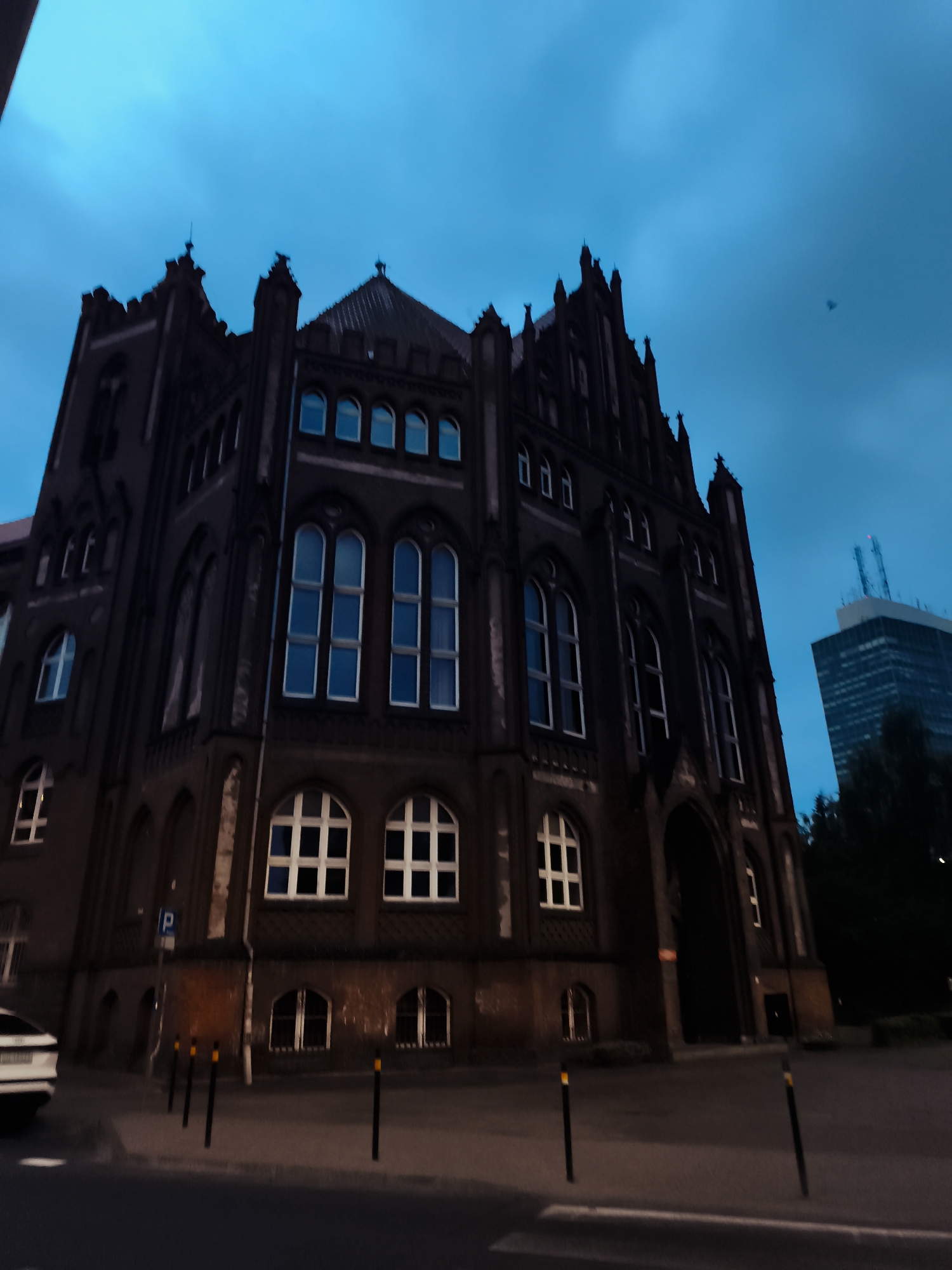 The image depicts the historic building of the former Main Post Office in Gdańsk, Poland, at dusk. The imposing, dark brick structure features Gothic Revival architecture with pointed arch windows and intricate detailing. The sky above is a deep, twilight blue, hinting at the transition from day to night. To the right of the main building, a modern skyscraper with a prominent antenna rises, creating a striking contrast between old and new urban architecture. The foreground shows a street with a few bollards and a portion of a white car. A parking sign is visible near the main building. The scene conveys a sense of historical grandeur juxtaposed with contemporary urban development.