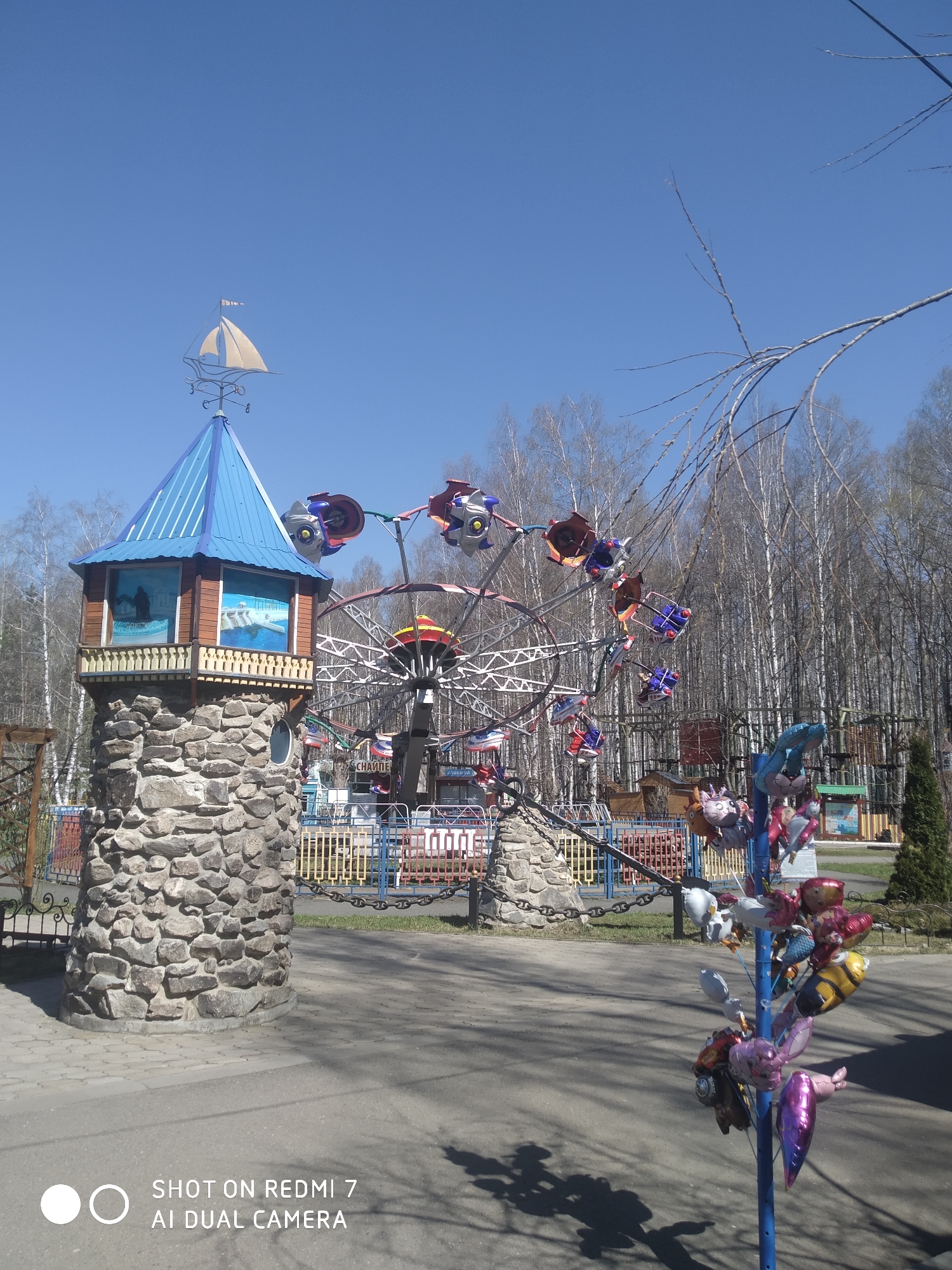 The image shows a clear, sunny day at a park in Chaikovsky, Russia. The foreground features a stone structure resembling a castle tower with a blue roof and a sailboat weathervane. A children's amusement park ride with seats shaped like airplanes is visible behind the tower. A post displaying various character-shaped balloons stands to the right of the tower. Bare trees fill the background, indicating early spring or late autumn. The ground is paved, with shadows cast by the structures and trees. A partial view of an inscription that reads "SHOT ON REDMI 7 AI DUAL CAMERA" is at the bottom of the image.