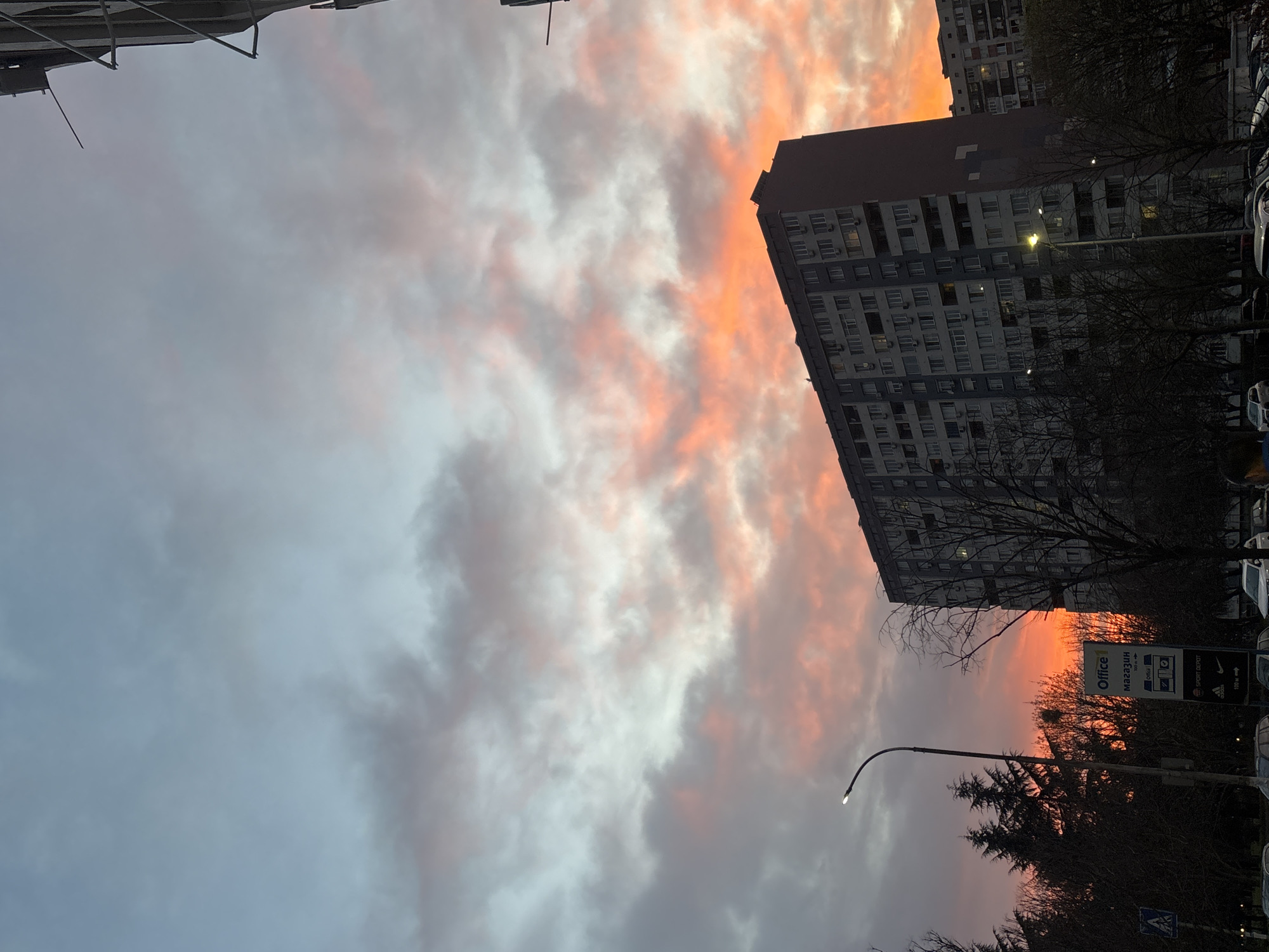 Captured at a low angle, the image showcases a dramatic sky at dusk or dawn, where clouds are illuminated with a vivid orange glow, contrasting with areas of grey. This creates a stark visual juxtaposition. A multi-story building is visible on the right, its structure rectangular and its windows appearing dark. Bare trees and a streetlight are positioned in front of the building, adding depth to the scene. Several parked vehicles are lined up along the street. A sign is placed near the streetlight, displaying the words "Office 1 магазини". The location is Burgas, Bulgaria, based on the Cyrillic writing "магазини" on the sign.