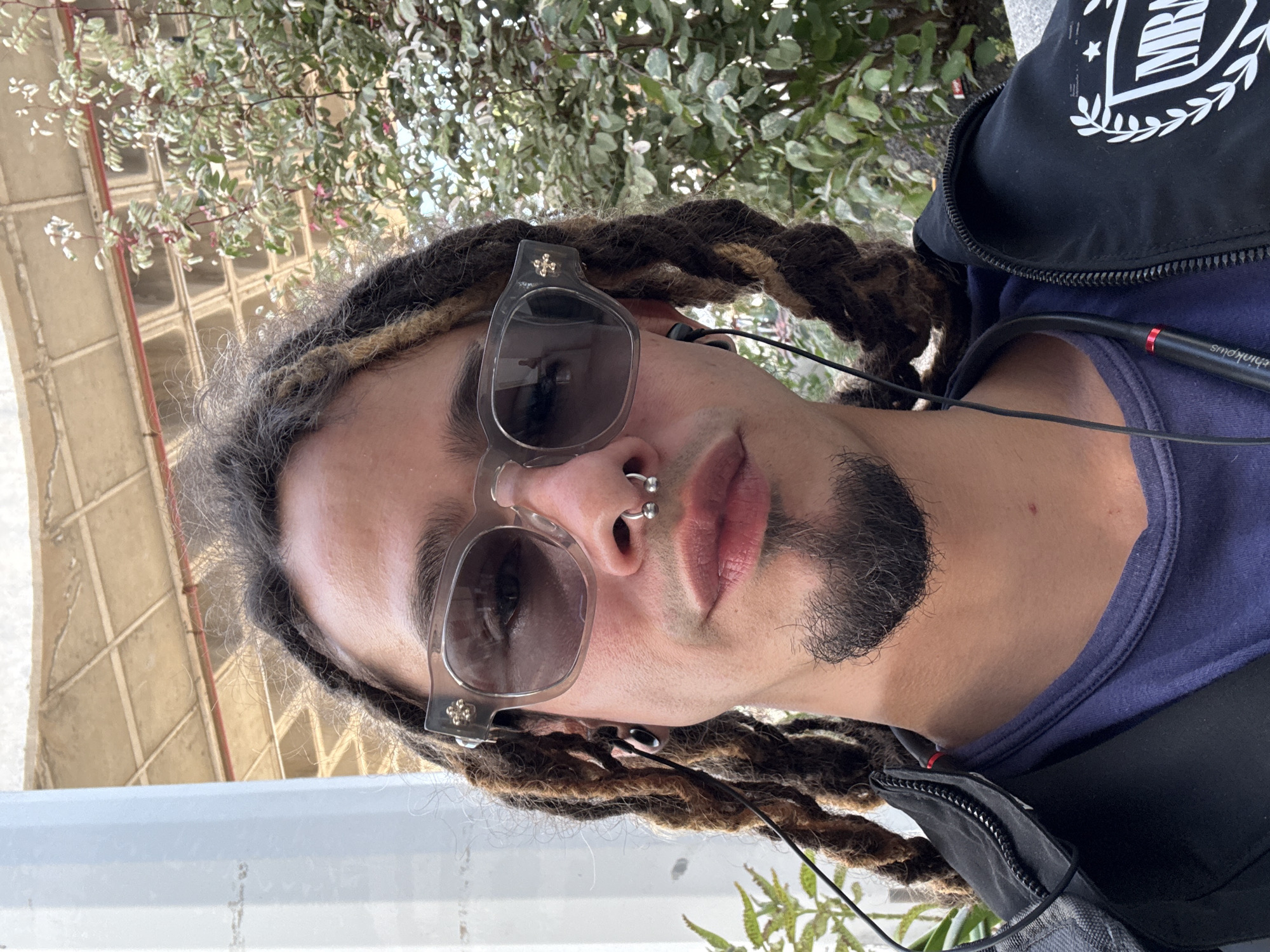 A medium shot depicts a male with dreadlocks and sunglasses. He has a nose ring and a short beard. He is wearing a navy blue t-shirt under a black zip-up jacket. Headphones are visible. Behind him are plants and what appears to be a beige architectural structure. While the location is specified as Montaña Roja, Spain, this cannot be confirmed from the visual information alone.