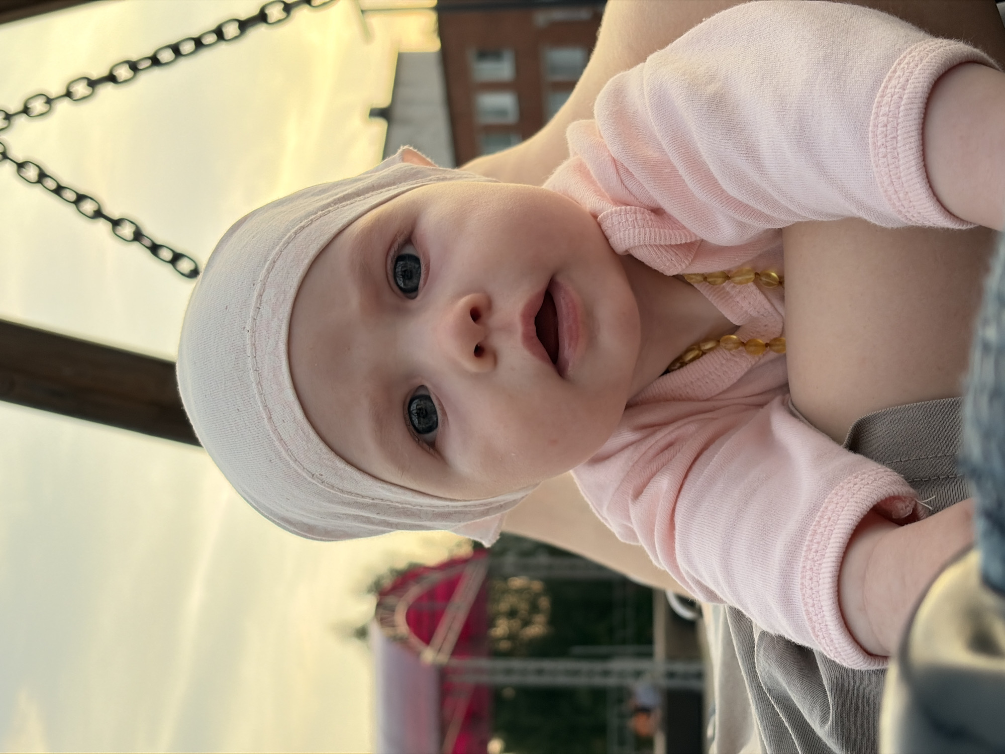An infant wearing a pale pink onesie and a matching headscarf is lying on their stomach, looking at the camera. The baby has dark blue eyes and a surprised expression with their mouth slightly open. A strand of amber beads is visible around the infant's neck. The background is blurred, showing a sunset sky with some clouds, a red-roofed structure, buildings, and a swing set. The image was taken outdoors, possibly in a park or playground setting in Noviy, Russia.