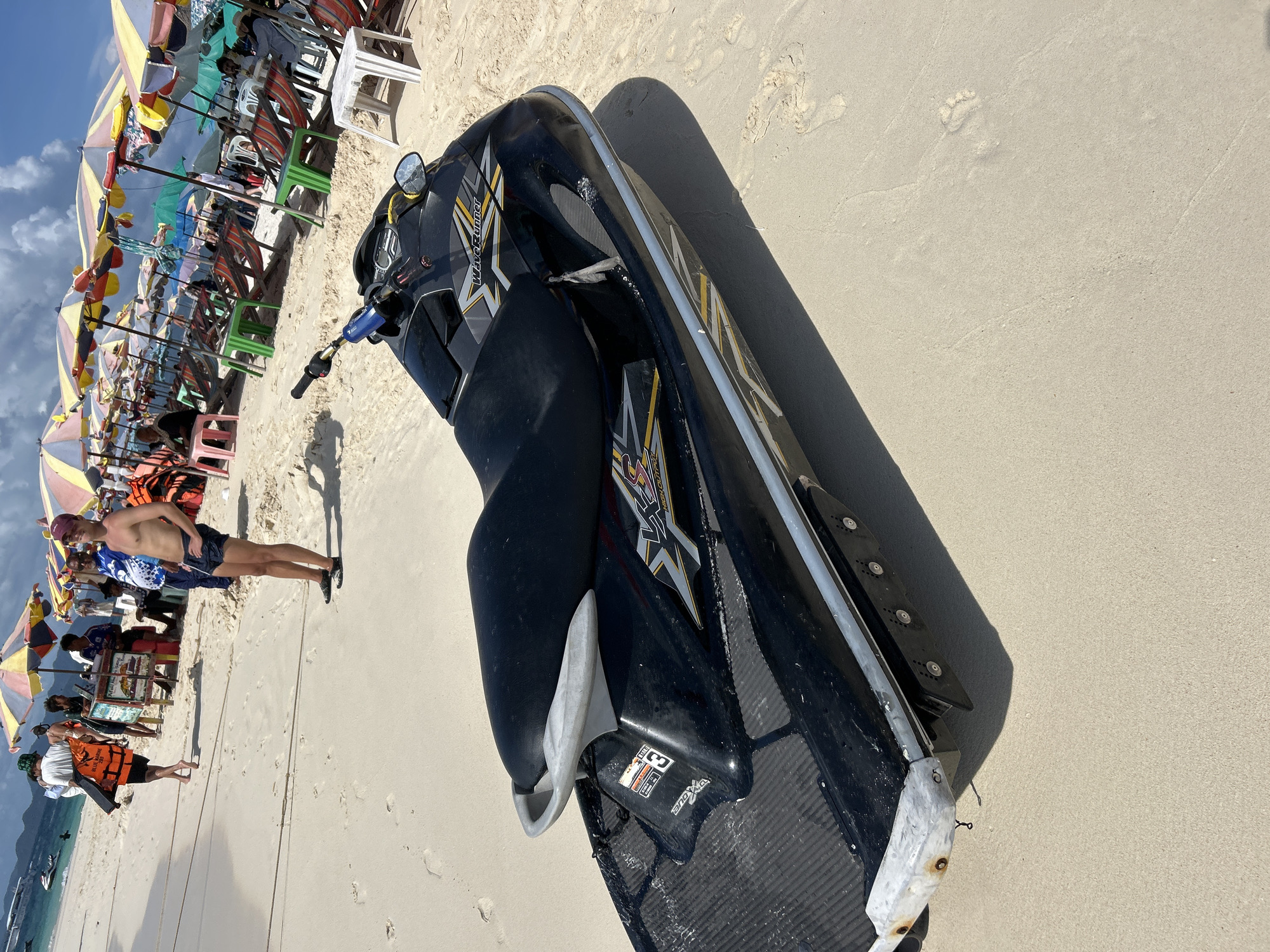 On a bright, sandy beach at Phru Nai, Thailand, a black Yamaha WaveRunner jet ski is parked in the foreground. The jet ski is positioned at an angle, with its rear facing the viewer. Its seat is black and has a silver grab handle. Decals with yellow and silver accents are visible on the body of the jet ski. In the background, a line of colorful beach umbrellas and lounge chairs is set up along the shore. Several people are present, some walking on the beach and others sitting in the chairs. One person is wearing an orange life vest. The sky is partly cloudy.