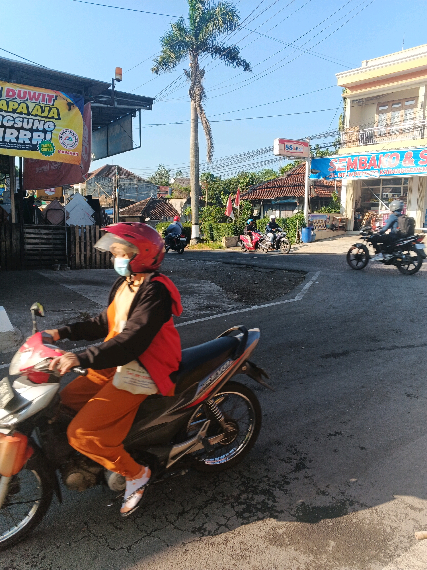 This image depicts a street scene in the city of Semarang, Indonesia. In the foreground, a person wearing a red helmet, a light blue face mask, and orange pants is riding a red and white motorcycle. The rider is wearing a black jacket over an orange shirt and white socks with an orange pattern. The motorcycle is moving on a paved road with visible cracks.
In the background, several buildings are visible. To the left, there is a building with a banner that reads "DUWIT APA AJA LANGSUNG RRRR!" and a smaller banner with "MAPAGAN". A tall palm tree is prominent in the middle ground. To the right, there is a multi-story building with a sign that says "88 Mart". Further back, another building with a blue and white sign reads "SEMBAKO & S". Multiple other motorcycles and riders are also present in the scene, some moving and some parked. The sky is clear and blue, indicating daylight. Electrical wires crisscross above the street.