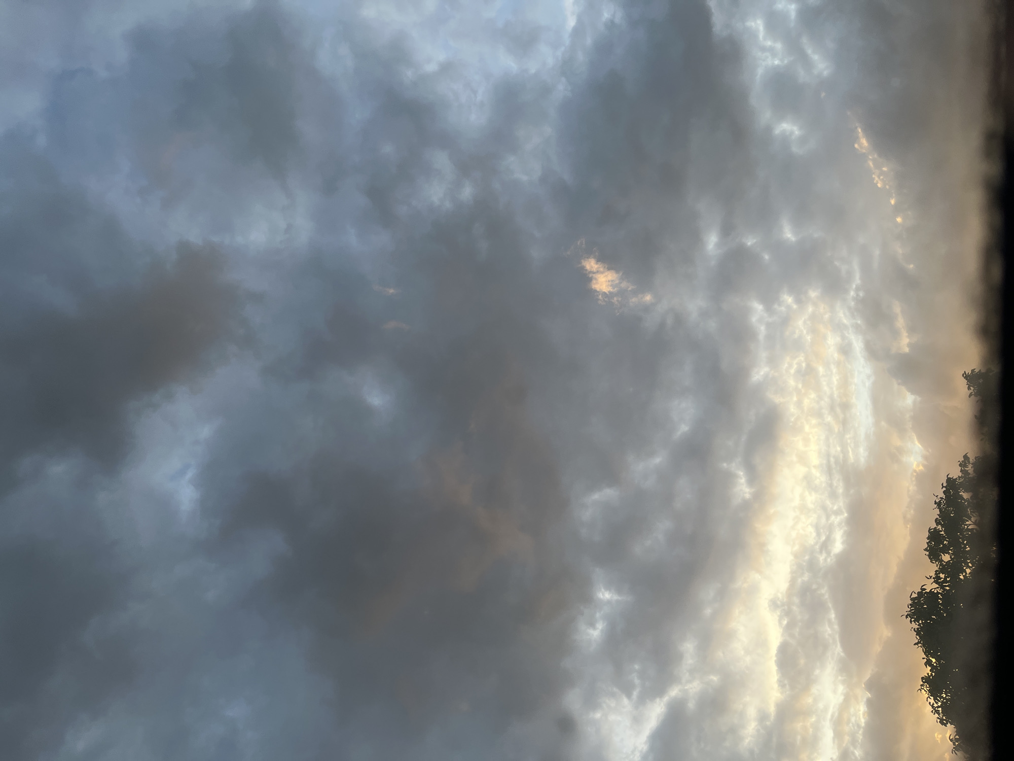 The image depicts a sky filled with dramatic clouds at dusk. The clouds are a mix of dark gray and lighter, wispy formations, with hints of orange and yellow light breaking through in some areas. The lower portion of the image shows the silhouette of trees and a dark horizon line, suggesting a landscape view. The overall impression is of an impending storm or the aftermath of one, with the last light of the day struggling to penetrate the dense cloud cover. The scene is set in Mondercange, Luxembourg, though no specific landmarks are visible in this atmospheric sky view.