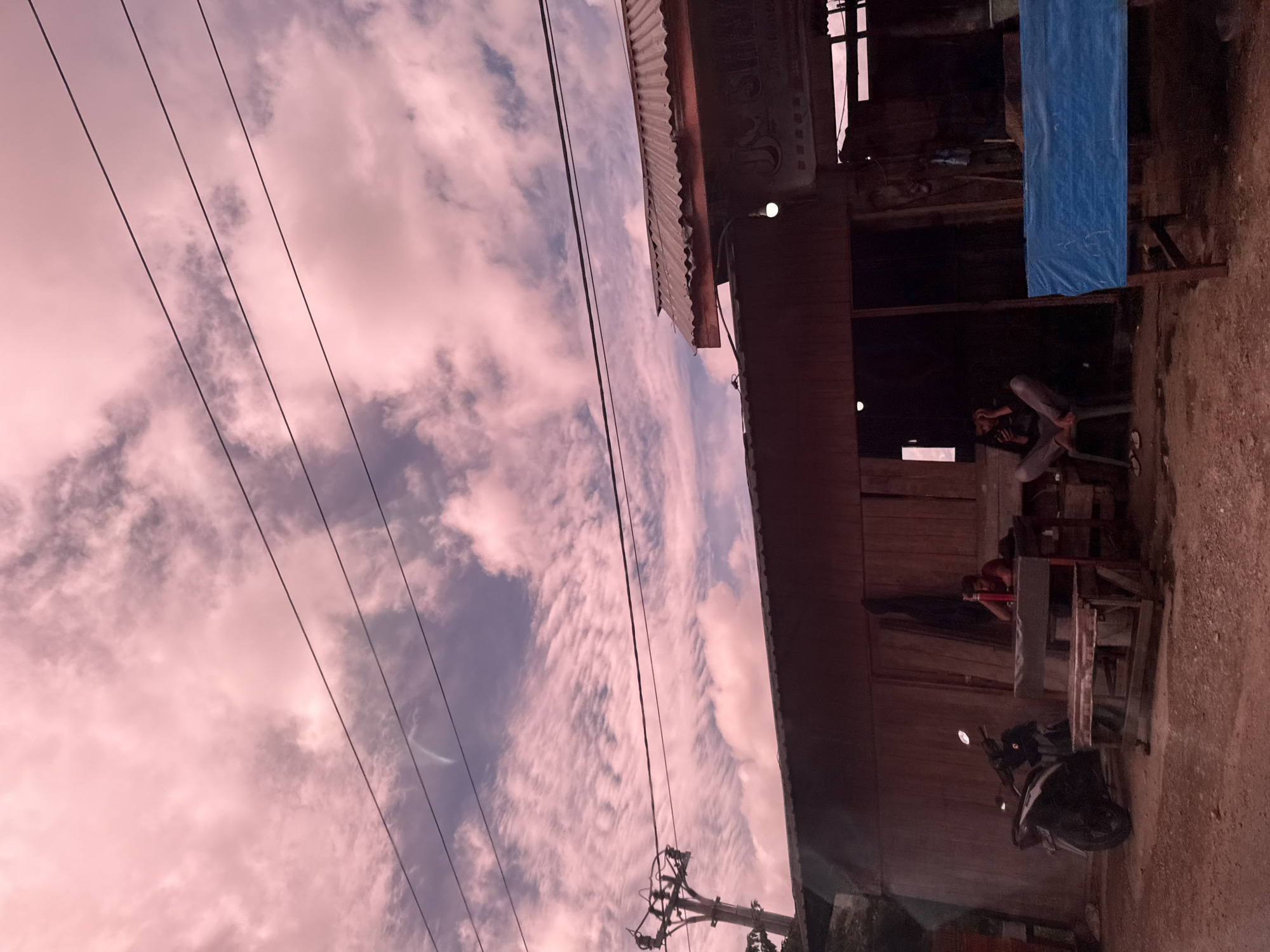 The image depicts an outdoor scene in Buengbakjok, Indonesia, under a pink-tinged sky filled with clouds. In the foreground, a wooden building with a corrugated metal roof stands. To the left of the building, a motorcycle is parked. In front of the building, two men are seated. One man, wearing a red shirt, sits at a wooden table, leaning on it with his arm. Another man, wearing a dark shirt and grey pants, sits on a chair to the right, looking at his phone. A blue tarp covers a table or structure to the far right. Power lines stretch across the sky, supported by a utility pole visible on the left. The ground appears to be a dirt and gravel surface.