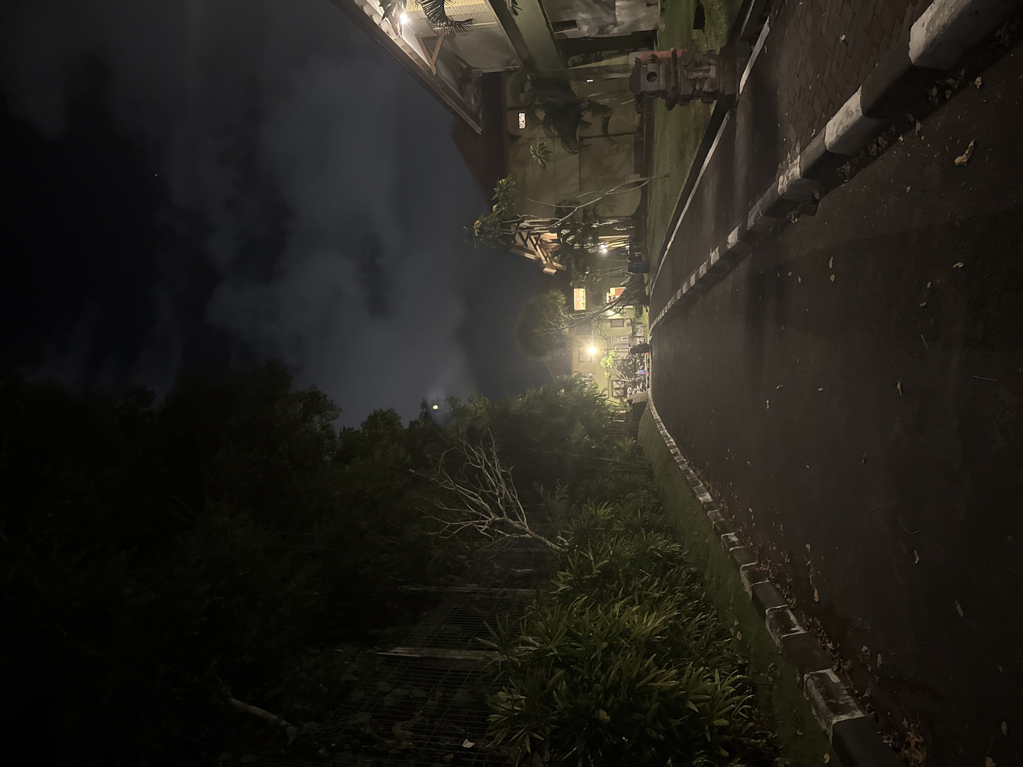 The image depicts a paved pathway at night in Kuta Selatan, Indonesia, illuminated by artificial lights. The pathway is bordered on the left by dense, dark green foliage and a wire fence, and on the right by a building with warm, lit windows and a textured facade. The sky above is dark with scattered clouds and a visible moon. The pathway itself is asphalt, with a concrete curb that is partially painted black and white. Fallen leaves are scattered on the pathway and the curb. In the distance, along the pathway, are more buildings and trees, also lit by external lighting, suggesting a resort or residential area. There are no people clearly visible, but the presence of a few distant figures can be inferred by faint lights or shadows. The overall atmosphere is tranquil and nocturnal.