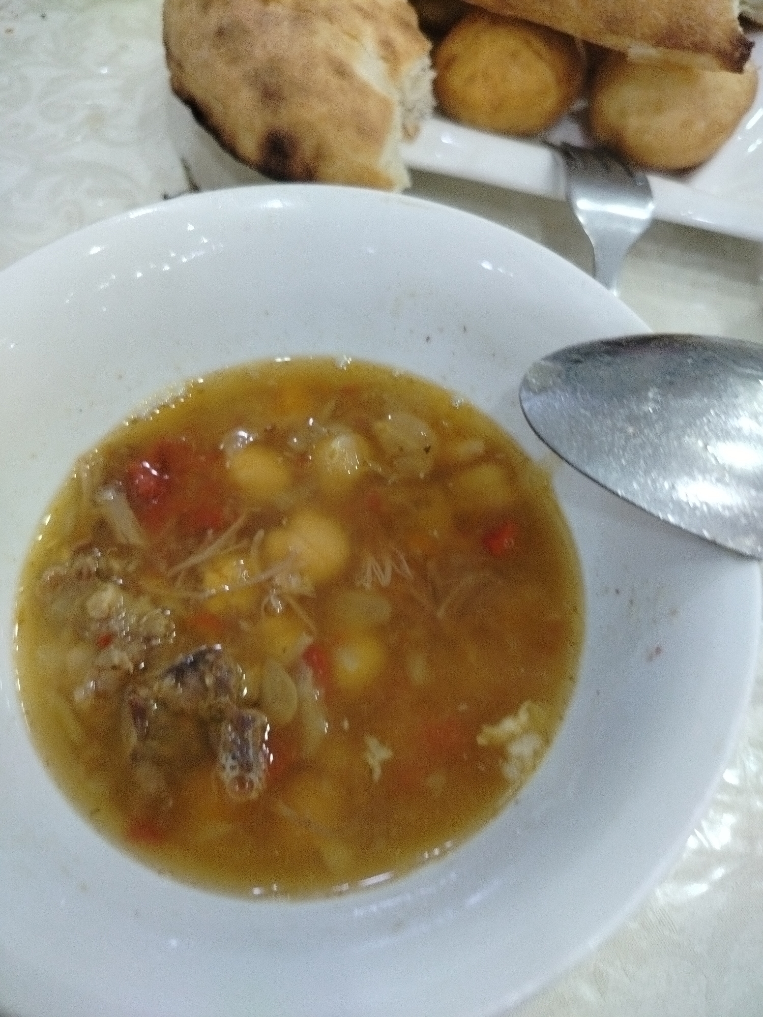A close-up, slightly blurry shot shows a bowl of light brown stew with visible pieces of meat, vegetables, and what appear to be chickpeas. A silver spoon rests on the right edge of the bowl, its reflection glinting. In the background, a plate holds pieces of bread and small, golden-brown fried items, possibly falafel or small pastries. The setting appears to be a casual dining environment, possibly a restaurant or home kitchen in the city of Aqsuat, Kazakhstan, given the context. The lighting is soft, creating a warm and inviting atmosphere, despite the image's lack of sharp detail.