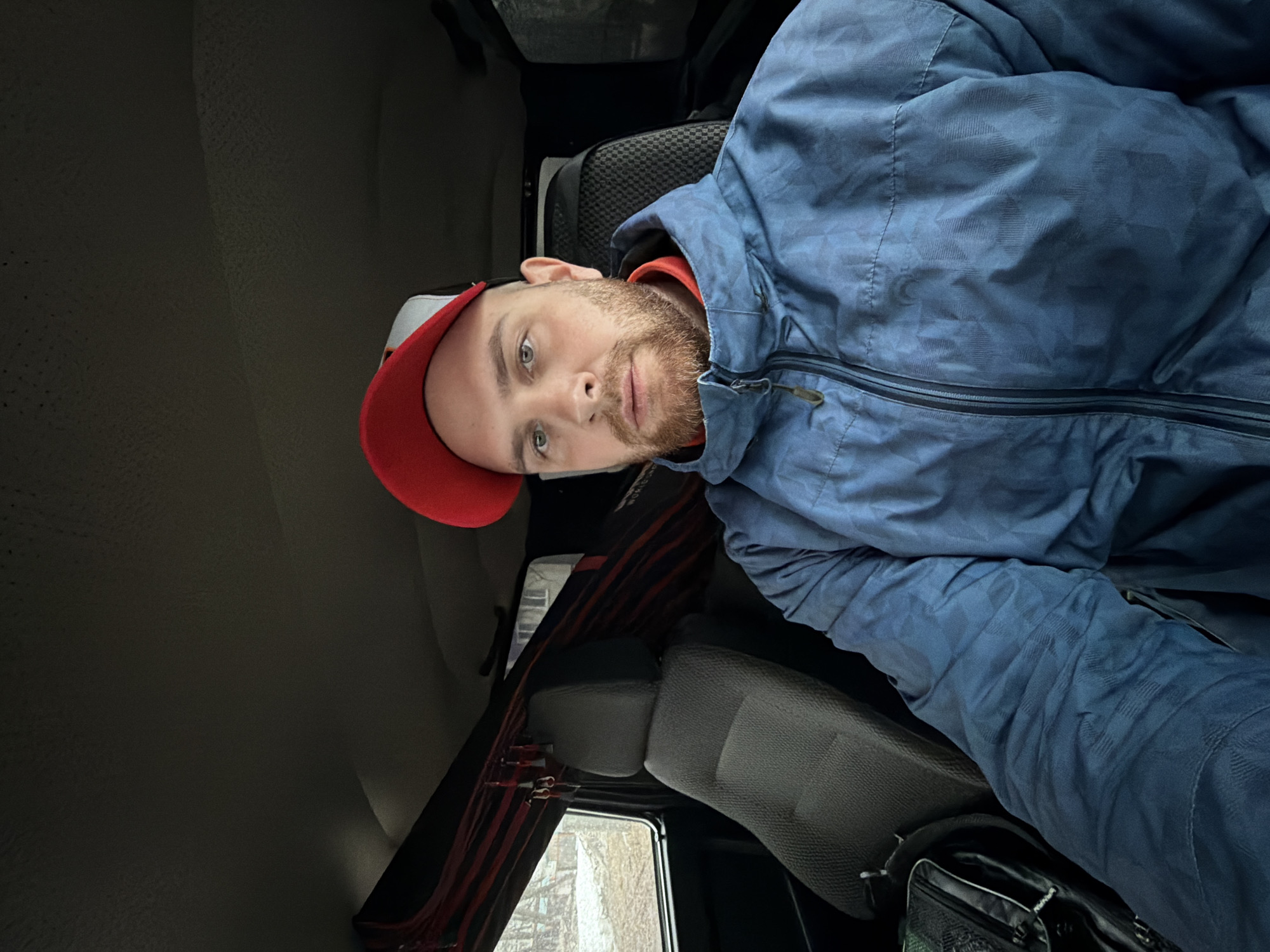 The image shows a man with a beard and a red baseball cap, wearing a blue jacket, seated inside a vehicle. The interior of the vehicle is visible, including the seats, window, and the roof. Through the window, a blurred view of the outside suggests an urban environment. Given the prompt, this is likely a scene within the city of Chaikovsky, Russia.