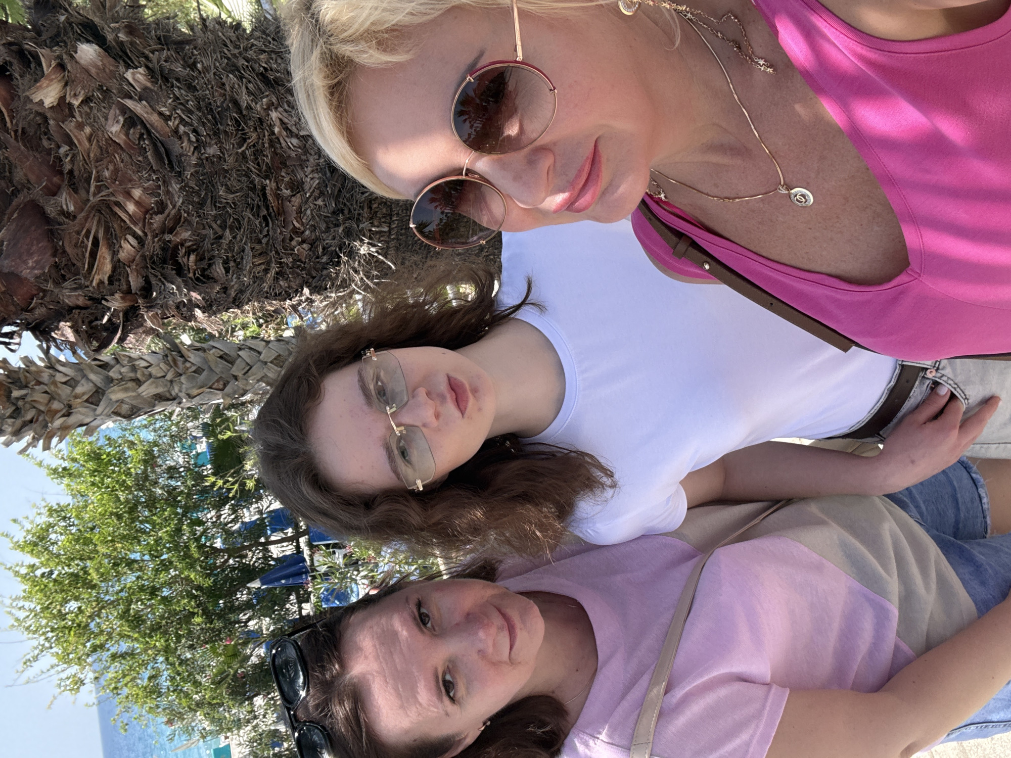 The image depicts three individuals in an outdoor setting. A woman with blonde hair, wearing pink sunglasses and a pink top, is positioned at the top of the frame. Below her is a younger individual with brown hair, wearing glasses and a white T-shirt. At the bottom is another woman with dark hair, wearing sunglasses and a light purple and beige top. They appear to be in close proximity, possibly taking a selfie.
The environment includes a palm tree trunk and foliage in the background, with a glimpse of the sea and beach umbrellas. The location appears to be a coastal area, city Unknown, Unknown.