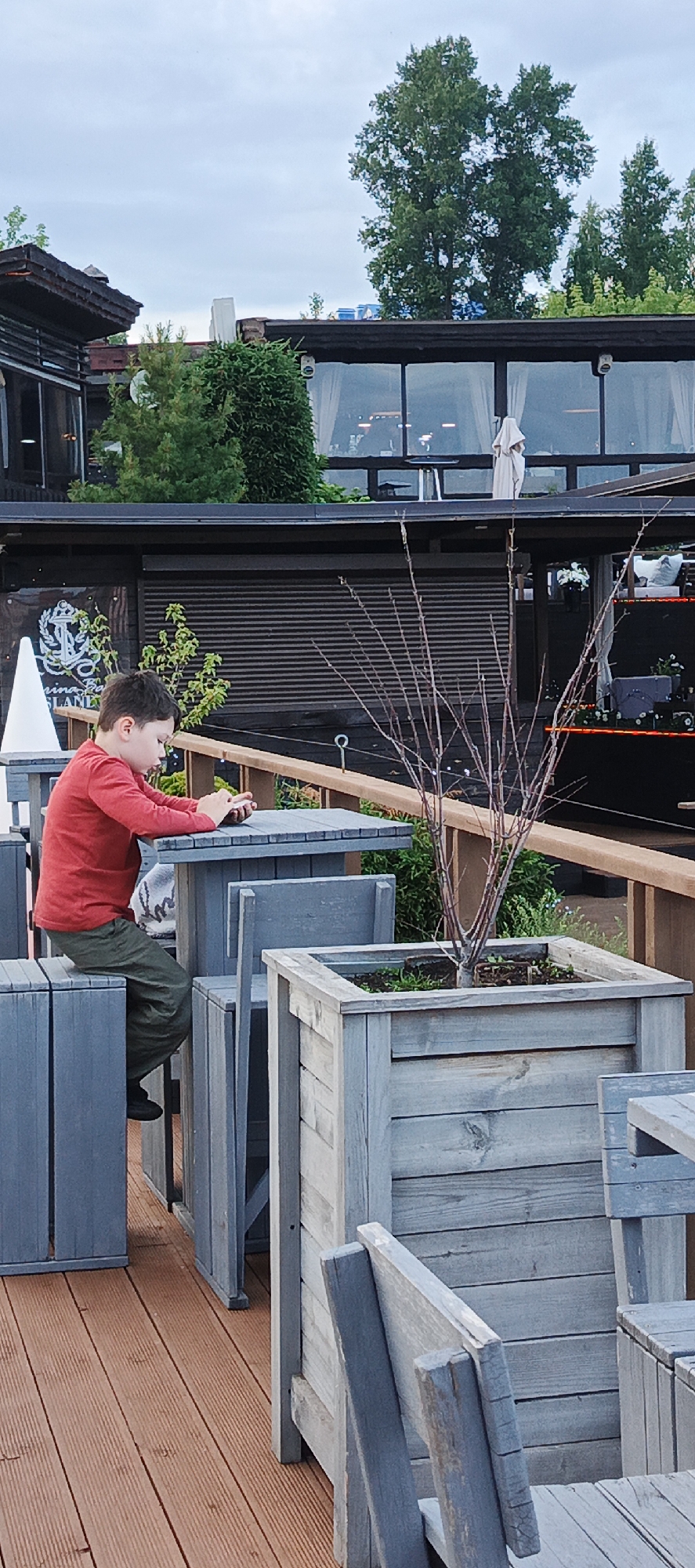 A young boy in a red long-sleeved shirt and green pants is sitting at a gray wooden table outdoors, looking down at his phone. He is on a wooden deck, which is a light brown color. Behind him is a gray wooden planter with a small bare tree. In the background, there is a building with large windows and a dark roof. The sky is overcast. The location is Votkinsk, Russia, specifically the restaurant "Marina Bay."