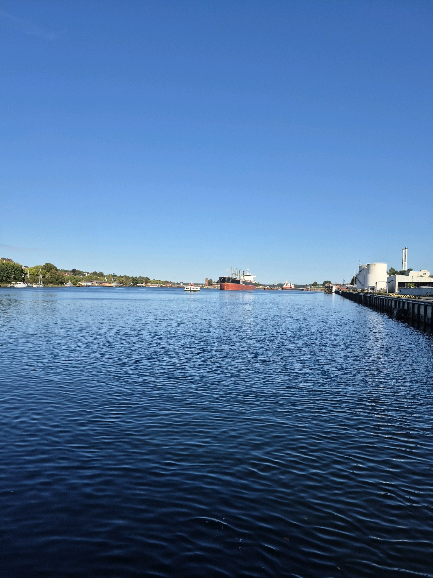 This image depicts a wide shot of the Kiel Fjord in Kiel, Germany, on a clear, sunny day. The vast, deep blue water of the fjord dominates the foreground, with gentle ripples creating a textured surface.
In the middle ground, on the left, a shoreline is visible with lush green trees and scattered buildings, suggesting a residential or park area. Several sailboats are anchored or moored along this side. A small white passenger ferry is navigating the water.
The right side of the middle ground shows an industrial or port area. A large, red-hulled cargo ship is prominently docked or anchored in the distance on the left side of the frame. Closer to the right, there are white industrial buildings, including large cylindrical storage tanks and a tall chimney stack. A long pier or dock extends into the water, lined with bollards and what appear to be large black fenders. The pier is also a walkway or road.
The background is a clear, bright blue sky, with no clouds visible. The overall impression is one of a calm, active maritime environment in Kiel.