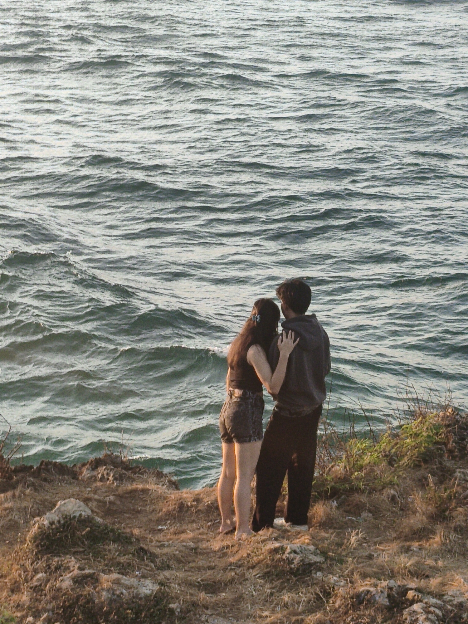 An over-the-shoulder, medium shot captures a young couple standing on a rocky cliff overlooking the ocean. The man, on the right, wears a dark grey hoodie and black pants, his arm around the woman. The woman, on the left, has long brown hair with a blue clip and wears a black tank top and denim shorts. Her arm is resting on the man's chest. The sea, with its characteristic choppy waves in a greenish-blue hue, fills the background. The cliff face is covered in dry grasses and sparse vegetation. The scene is set during the golden hour, with the warm sunlight casting a soft glow on the landscape. The location is Kuta Selatan, Indonesia, likely near a coastal viewpoint or cliffside path.
