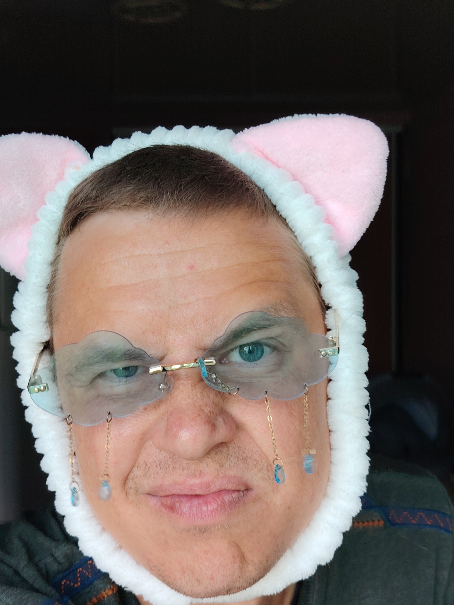 A head-and-shoulder portrait captures an adult male with fair skin and visible facial stubble. He is wearing a white, fluffy headband that features two soft pink, rounded cat ears positioned above his dark brown hair. His blue eyes are visible through light blue-tinted, cloud-shaped, rimless glasses. These glasses have a gold-toned bridge and temple arms, and are adorned with four small, light blue, teardrop-shaped beads suspended by delicate gold chains from the lower edges of the cloud lenses, with two beads on each side. The subject's expression is neutral, and he gazes directly towards the viewer. He is dressed in a dark green garment, possibly a sweater or shirt, which displays a patterned band with orange, blue, and brown geometric designs on his right shoulder. The background is uniformly dark, indistinct, and out of focus. There are no discernible elements within the image to indicate its specific location in the city of Chaikovsky, Russia, or any other geographical area.