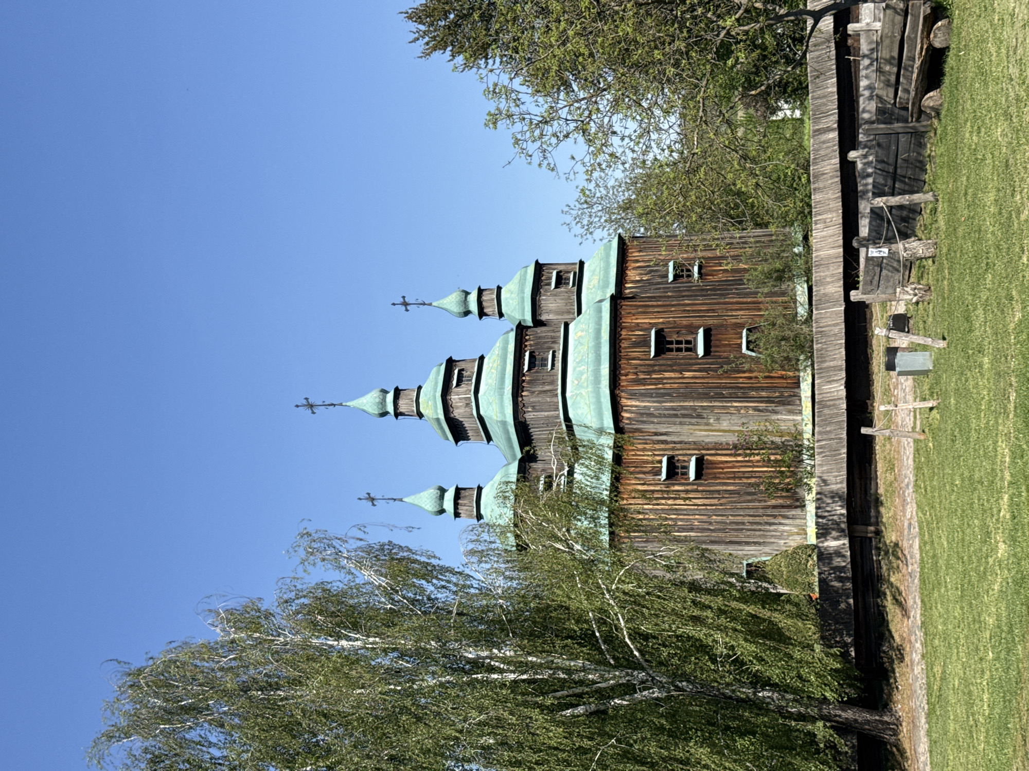 An exterior shot reveals a wooden church with three domes, likely the Church of Saint Paraskeva Piatnytsia, set against a clear blue sky. A birch tree with green leaves occupies the lower left of the frame. Adjacent to the church is a wooden fence and a grassy area. The church is located in Kyiv, Ukraine.
