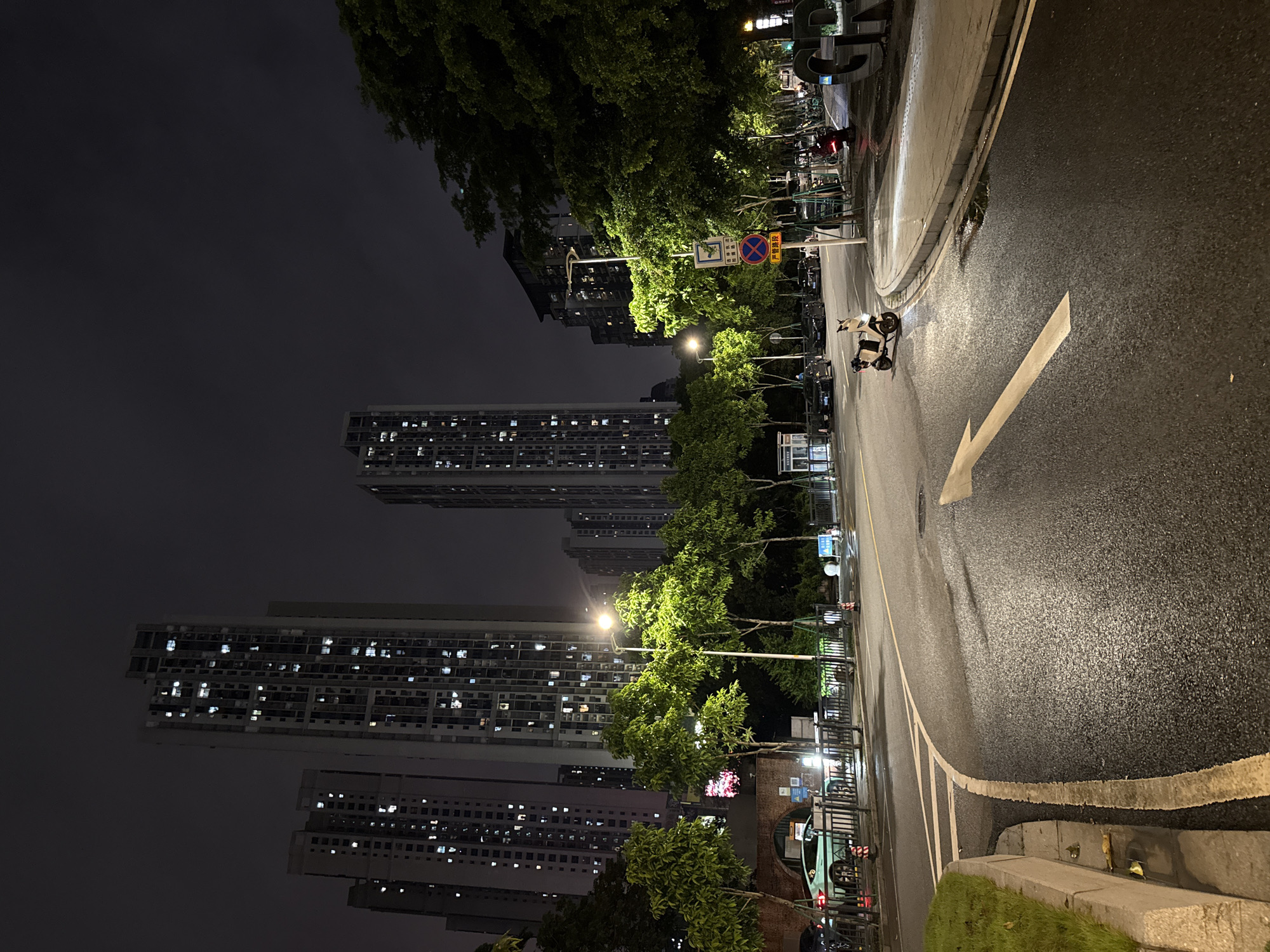 In Haizhu District, China, at night, a road curves to the right under a dark sky, illuminated by streetlights and building lights. A prominent white arrow painted on the wet asphalt indicates the direction of travel. A light-colored scooter with a person on it is stopped on the road to the right of the arrow. The background features tall, modern apartment buildings with many lit windows, indicating residential occupancy. Dense green trees line the street in the mid-ground, their leaves catching the light. A black fence separates the sidewalk from the road. Traffic signs, including a red circular sign with a diagonal bar and a blue square sign with a white symbol, are visible on poles along the sidewalk. Several cars are parked and driving on the street and sidewalk. A large, dark metal sculpture, possibly letters, is visible on the far right. The overall scene depicts a well-lit urban environment at night.