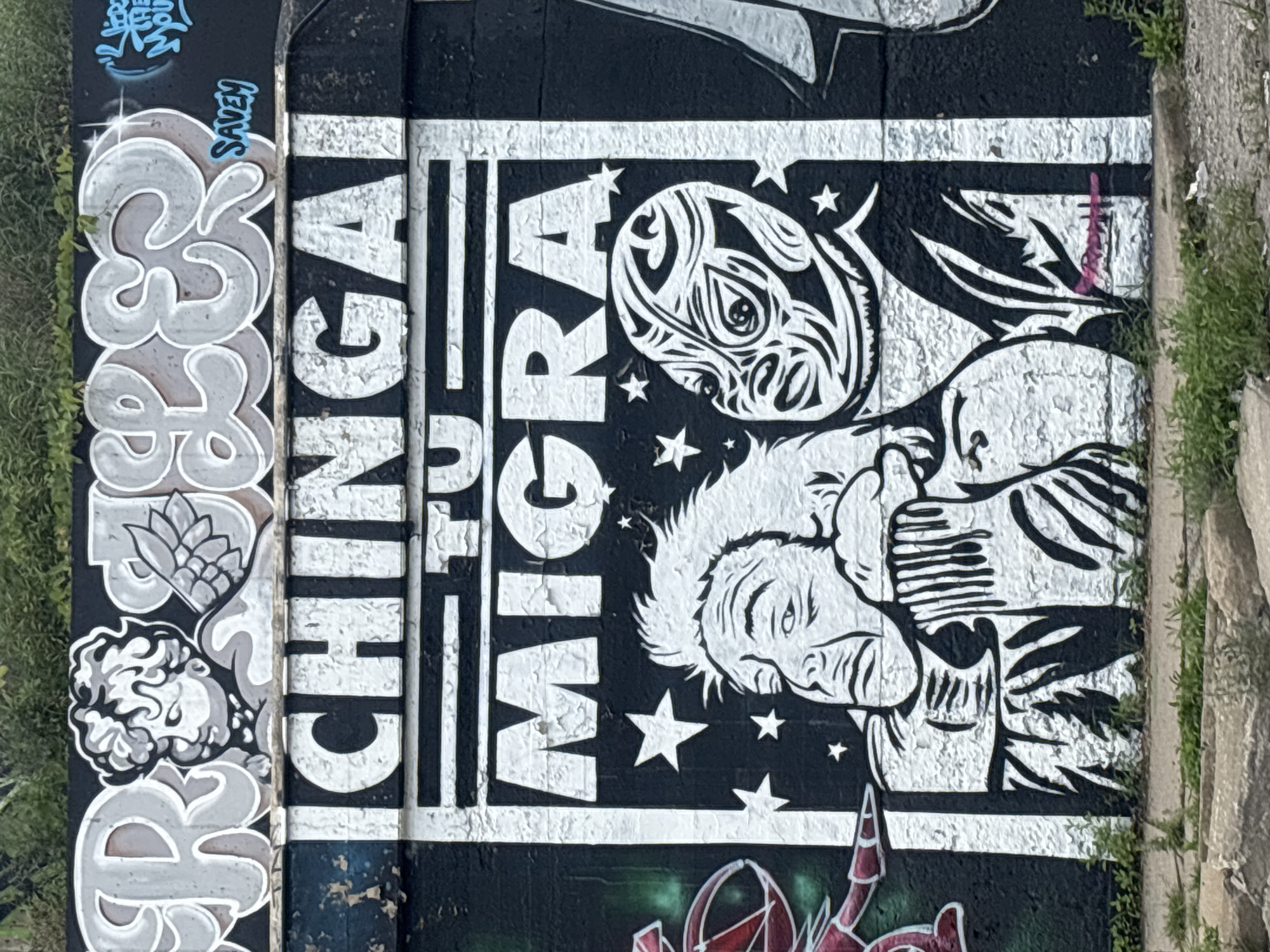 This is a photograph of graffiti art on a black wall in Chicago, Illinois, United States of America. The graffiti is predominantly black and white, with some grey and blue accents.
At the top of the wall, large grey bubble letters are painted, with a cherubic figure with wings positioned in the center. Below this, a large black rectangular panel contains white lettering that reads "ICHINGA TU MIGRA" in bold, block capitals. The text is stylized, with some letters outlined in black and others solid white.
Within the black panel, there is a graphic depiction of two figures. The figure on the left has a pained expression, with its head being held by the figure on the right. The figure on the right wears a mask with a tribal or wrestling design, featuring black and white patterns. Several white stars are scattered around the figures.
To the left of the central panel, a portion of another graffiti piece is visible, featuring red and green colors and a stylized figure. To the far right, some blue spray paint graffiti is also present.
The wall appears weathered, with peeling paint and a rough texture. The bottom of the image shows the edge of a concrete surface and some overgrown grass and weeds.