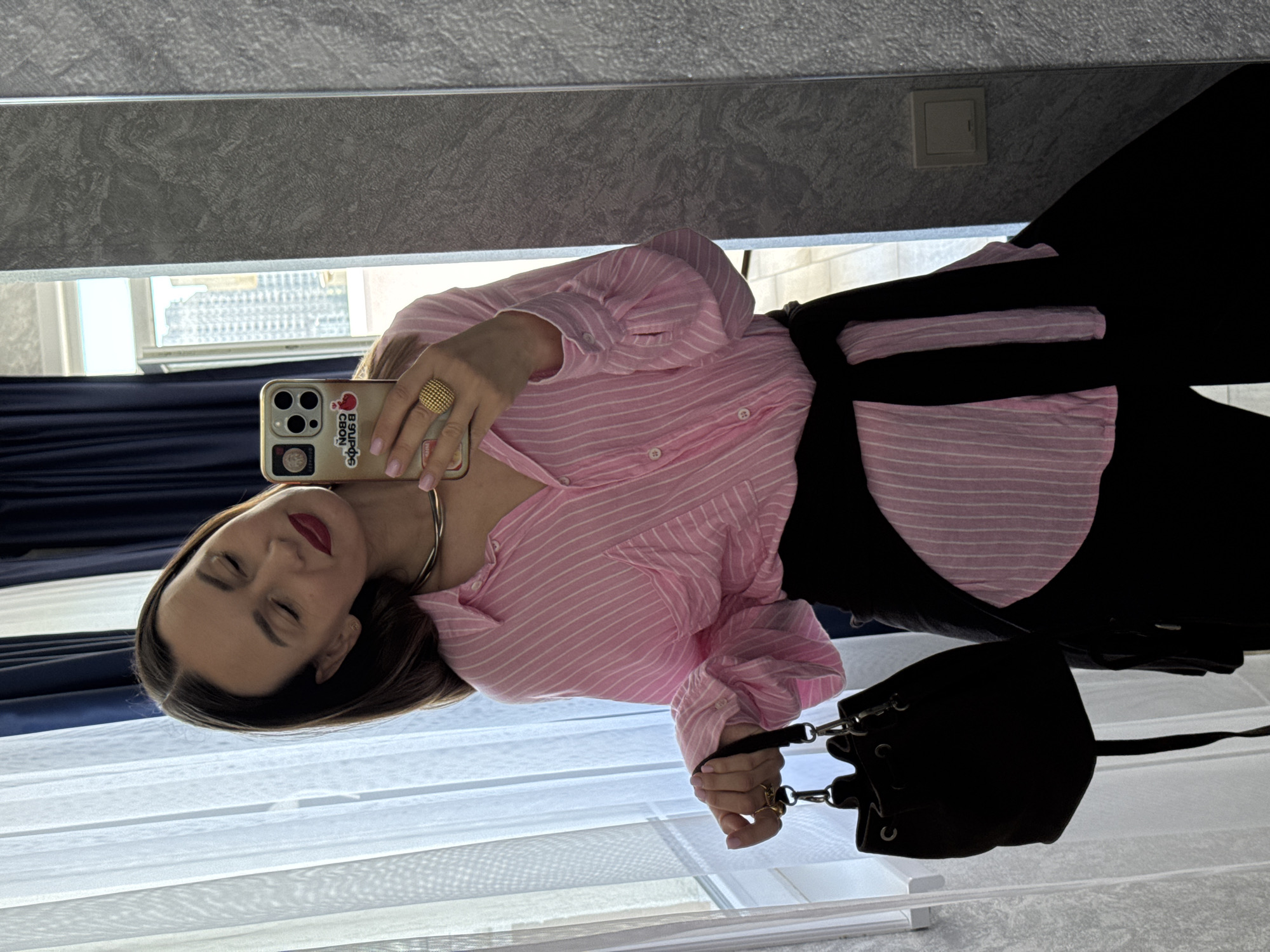 This rotated mirror selfie captures a young woman in an indoor setting in Sochi, Russia, likely a room with a window. The image is oriented 90 degrees counter-clockwise.
The woman is the central subject, posing with her eyes closed and a subtle smile on her face, accented by red lipstick. She has straight, dark brown hair. She is dressed in a pink and white striped button-down shirt, partially unbuttoned to reveal a silver-toned choker necklace. Her lower attire appears to be dark, possibly black trousers or a skirt.
In her right hand, she holds a light-colored iPhone with a multi-lens camera system. The phone case is adorned with several stickers, including one with text in Cyrillic, "В БЮРО СВОЙ" (V BYURO SVOY, meaning "My own in the office" or "Own in the bureau"). On her right ring finger, she wears a prominent, spherical gold ring with a textured, beaded design. Her fingernails are painted a light pink. A small black handbag with silver-toned hardware is visible, held by her left hand.
The background, visible in the mirror's reflection, shows white translucent curtains or blinds covering a window, through which a building structure can be vaguely seen outside. A dark gray roller blind or shutter is partially lowered above the window. An electrical outlet or switch is present on a dark wall section. The lighting suggests it is daytime.