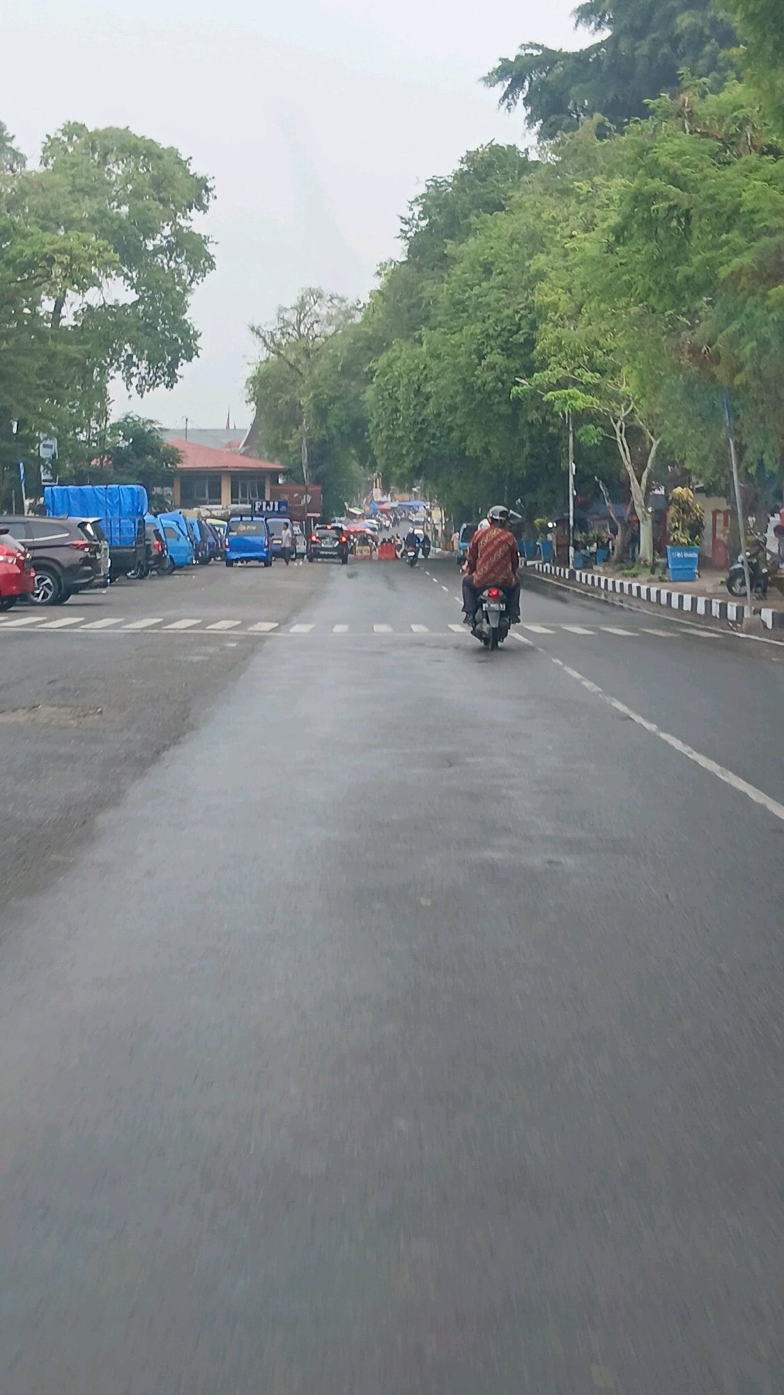 The image depicts a street scene in Batusangkar, Indonesia, on a cloudy day. In the foreground, a wet asphalt road stretches into the distance. A motorcycle carrying two passengers is moving away from the camera in the right lane. The passengers are wearing helmets, and the rider is wearing a patterned shirt.
Along the left side of the road, several vehicles are parked, including a red SUV, a dark-colored truck with a blue tarp, and several blue mini-vans or trucks. The buildings in the background on the left appear to be commercial establishments, with a building displaying the sign "FIJI". Trees line the right side of the road, with lush green foliage indicating a tropical or sub-tropical climate. Further down the road, traffic is visible, and there appear to be vendors or stalls set up on the right side. The overall impression is of a busy urban street.