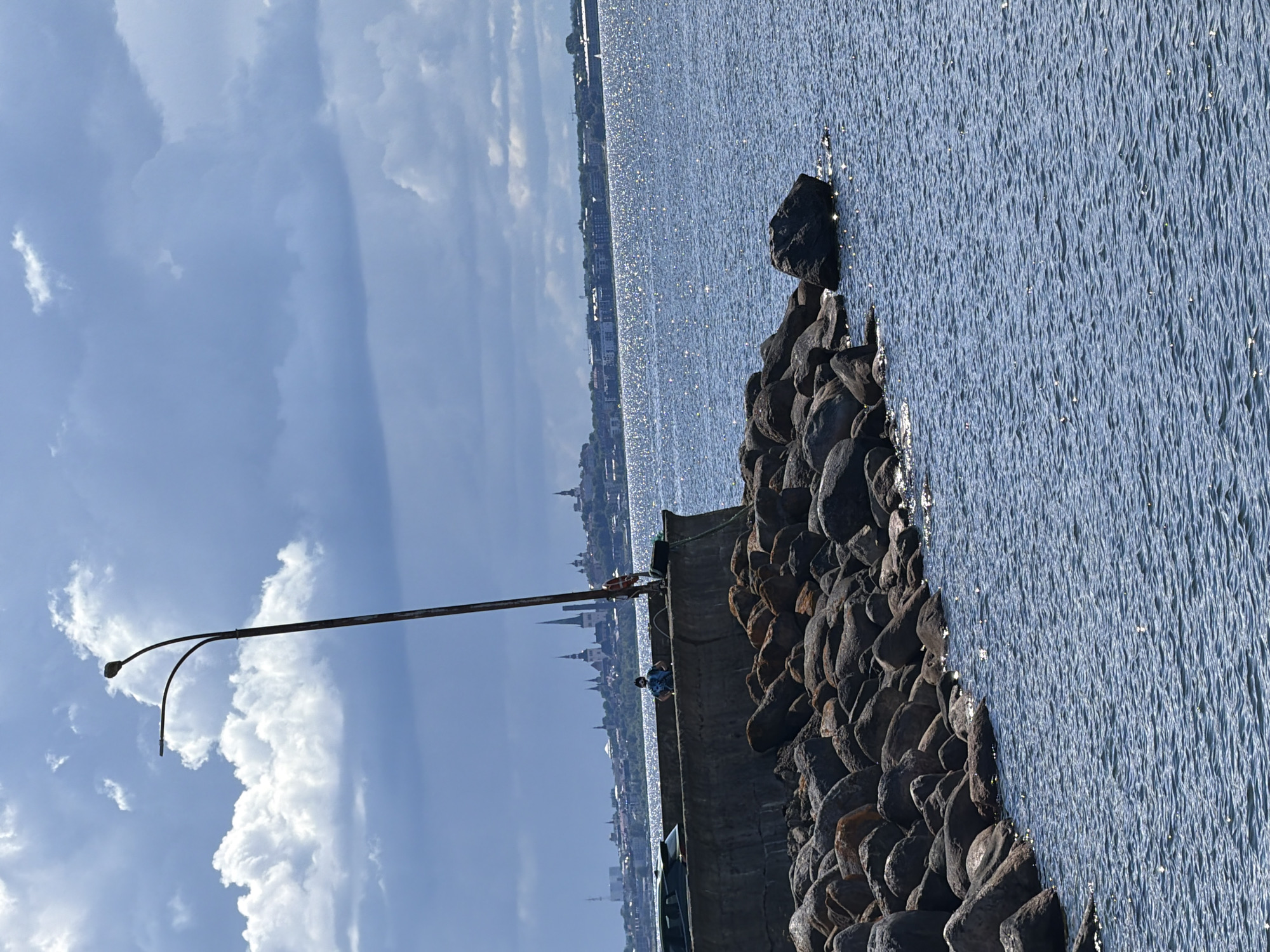 A full shot shows a man standing on a concrete pier with a rocky shoreline. The man is wearing a patterned shirt and dark pants. He is facing forward, with his hands on his hips. A street lamp with a curved arm extends upwards from the pier, positioned behind the man. A dark-colored car is parked on the pier to the left of the man. In the background, a city skyline with numerous spires is visible across the water, suggesting it is the city of Tallinn. The water is blue and choppy, with sunlight glinting off the surface. The sky is partly cloudy with dark gray clouds. This scene appears to be located in Viimsi, Estonia, likely near the coast offering a view of Tallinn.