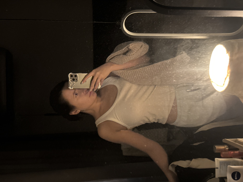 An adult female is depicted in a mirror selfie within a dimly lit indoor environment, identified as the city of Селекционер-2 Снт, Russia. She wears a white tank top and light-colored sweatpants. Her dark hair is pulled back. She holds a smartphone with a light-colored case featuring ear-like protrusions in her right hand, positioned to capture her reflection. A light-colored, ribbed textile, possibly a towel or scarf, hangs over her left forearm. The mirror surface exhibits visible particles and smudges. In the lower central foreground, a bright, warm-toned cylindrical light source illuminates the scene. A partial reflection of an arched light fixture is visible on the right. To the lower left, on a reflective surface, several personal care products are arranged, including a white tube labeled 'NIVEA'. The background beyond the reflection is largely dark.