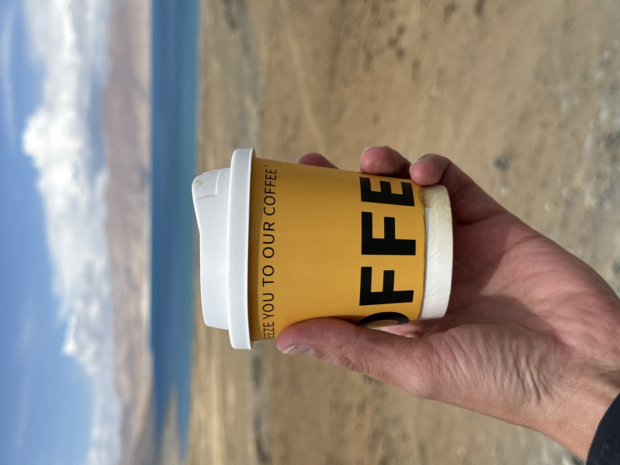 A hand holds a yellow paper coffee cup with black "OFF" letters and the phrase "FREEZE YOU TO OUR COFFEE" in front of a lake surrounded by arid land and mountains under a blue sky with white clouds. The location is Gyirong County, China.