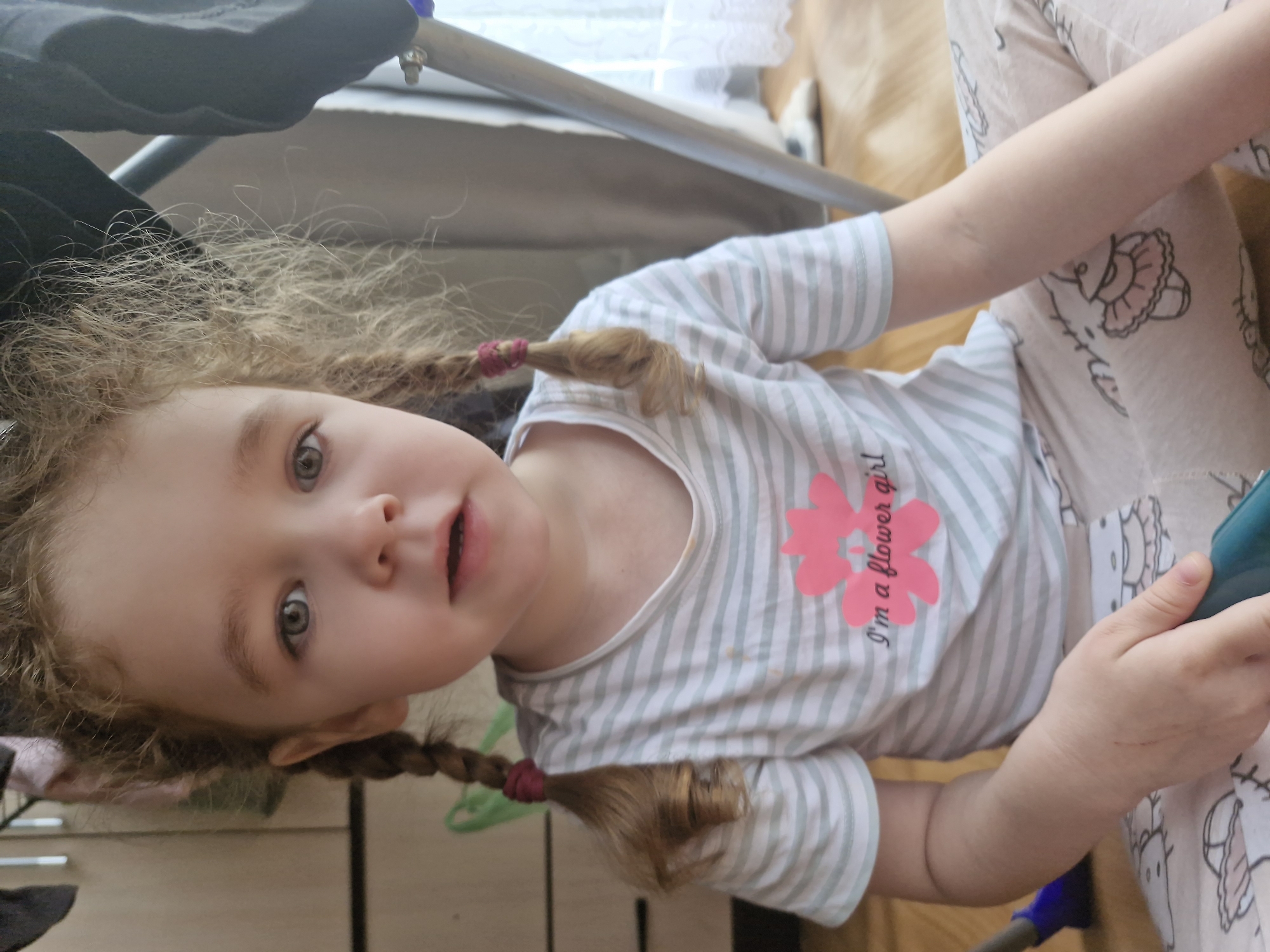 A young child with curly, light brown hair styled into two braids and a messy top knot sits on a wooden floor. The child is wearing a striped t-shirt with a pink flower graphic and the text "I'm a flower girl," and patterned pajama pants with cartoon designs. The child's eyes are open, looking directly at the camera. In the background, a wardrobe and part of a clothes rack are visible, along with a window with white curtains. The scene appears to be an indoor residential setting, likely a home, in the city of Марковский, Russia.