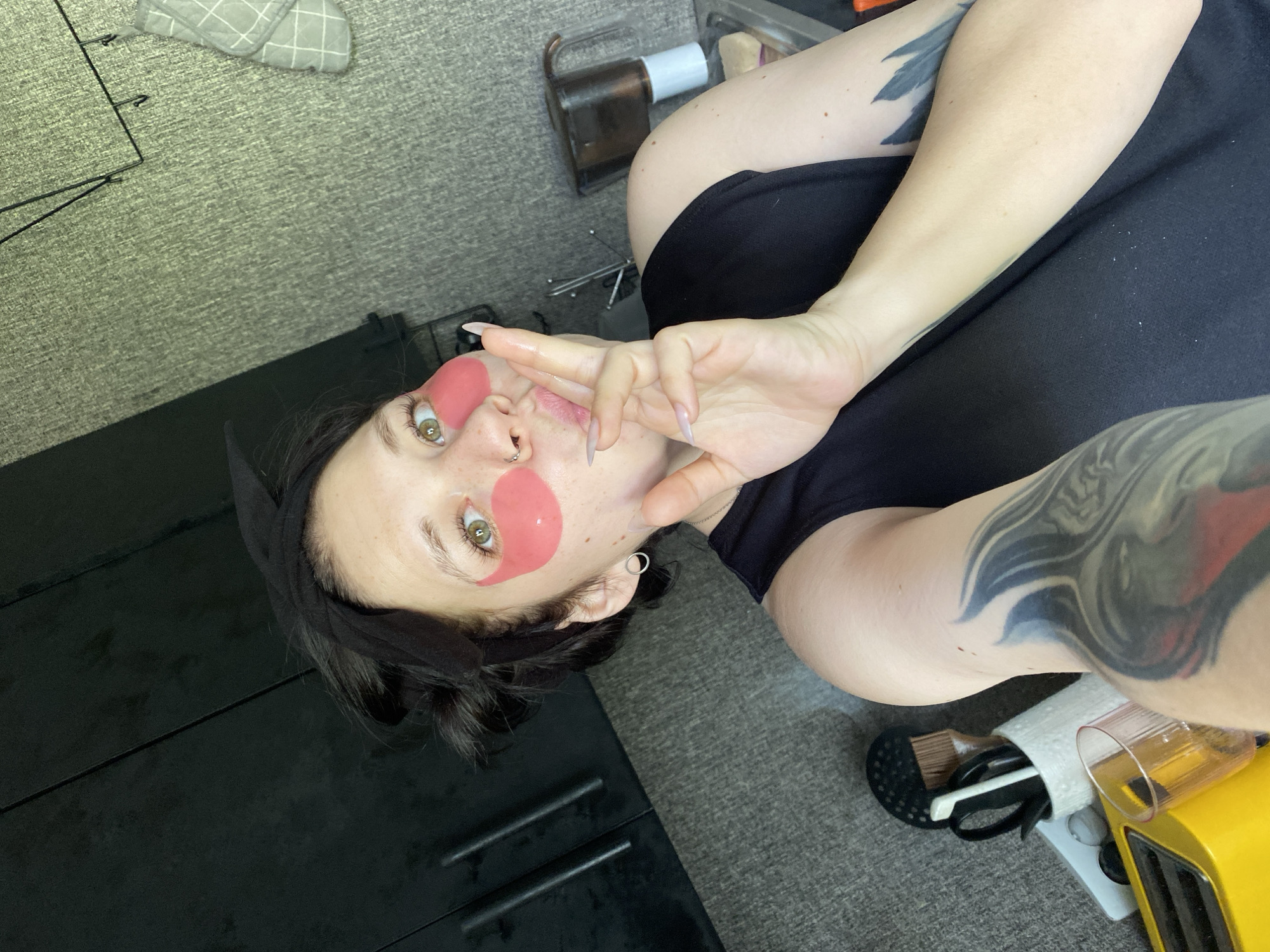 The image shows a person lying on what appears to be a gray carpeted floor. The person is wearing a black headband and has pink eye masks under their eyes. Their left hand is raised, forming a peace sign, with elongated, manicured nails. The person has visible tattoos on their arms. The setting includes a black cabinet with drawer handles to the left, a storage container with cosmetic items to the right, and other personal items scattered around. The image was not taken in the city of Chaikovsky, Russia.