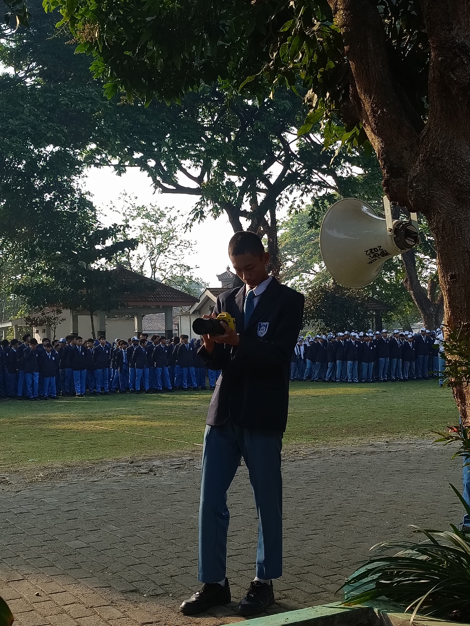 The image captures a group of students, presumably in uniform, standing in formation on a grassy field in what appears to be an outdoor school setting in Kediri City, Indonesia. The students, mostly male, are wearing dark blazers over light shirts and blue trousers. Some are also wearing white caps. In the foreground, a young male student is focused on a yellow camera he is holding, seemingly capturing the scene or an event. He is wearing a dark blazer, a light blue tie, and blue trousers. Behind the students, there is a building with a distinctive roof structure, possibly a pavilion or a small hall, and lush green trees provide shade. A large speaker is mounted on a tree to the right of the frame. The ground in the foreground is paved with interlocking bricks, transitioning to a gravel path. The overall atmosphere suggests a school event or assembly taking place outdoors in the morning or late afternoon, given the lighting.