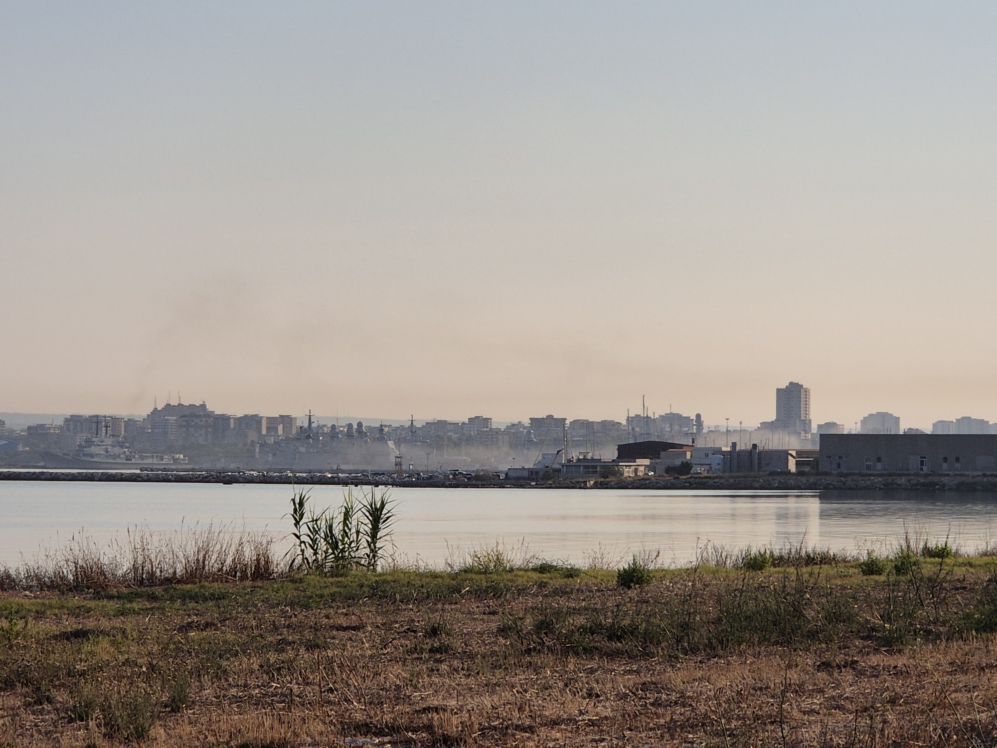 The image shows a wide, calm body of water in the foreground, with dry, brown grass and reeds along the bank. In the middle ground, there is a breakwater made of large stones, separating the foreground water from a harbor with several boats and ships. Beyond the harbor, the city skyline of Taranto, Italy, is visible. The buildings are a mix of older, multi-story structures and some more modern high-rises. The sky is clear and light, with a hazy quality, suggesting either early morning or late afternoon. Smoke plumes are visible in the distance, rising from within the city. The overall scene depicts a coastal urban environment.
