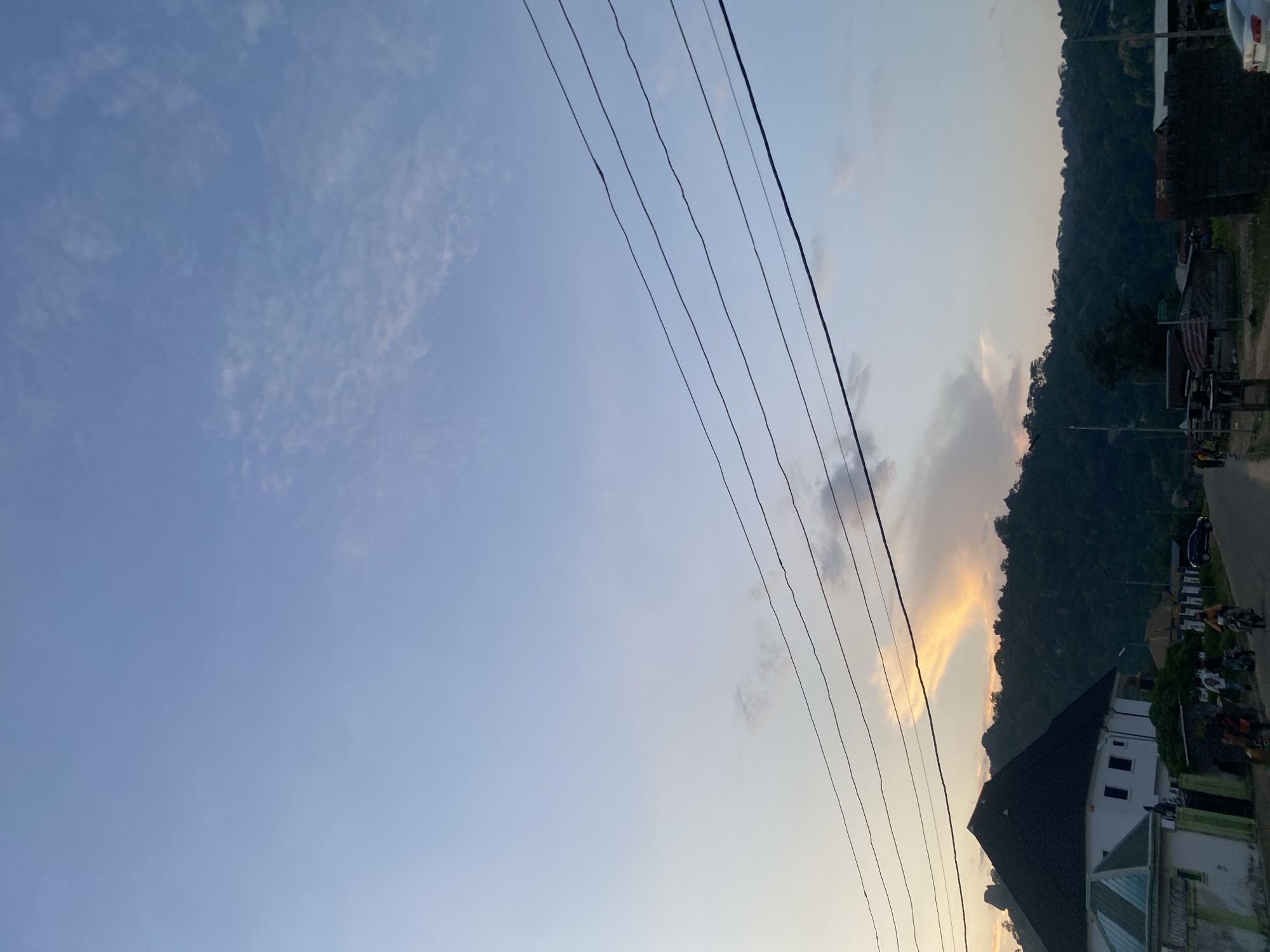 This image, captured in Enwan, Nigeria, depicts a late afternoon or early evening scene. The upper two-thirds of the frame are dominated by a pale blue sky, transitioning to a lighter, warmer hue near the horizon. Sparse, wispy white clouds are scattered across the sky, with a notable, elongated cloud formation above the left portion of the treeline exhibiting a distinct orange-yellow glow on its underside, indicative of a setting sun. Five dark, parallel power lines stretch horizontally across the middle of the sky.
The lower third of the image features a landscape comprising buildings, a road, and densely forested hills. In the lower-left foreground stands a prominent multi-story building with white walls and a dark, possibly black or dark grey, hip roof. To its immediate right, a smaller building with a light green-tiled roof and white walls is visible. Behind these structures, a range of dark green, tree-covered hills or mountains forms the backdrop, rising from the mid-ground.
A road traverses the lower section of the image, where several individuals and vehicles are present. At least two motorcycles, each with a rider, are visible on the left side of the road, moving towards the right. Other people are standing or walking along the roadside. A dark-colored SUV is parked on the right side of the road, and the front portion of a white car is partially visible at the extreme right edge of the frame. The overall lighting is soft and warm, characteristic of dusk.