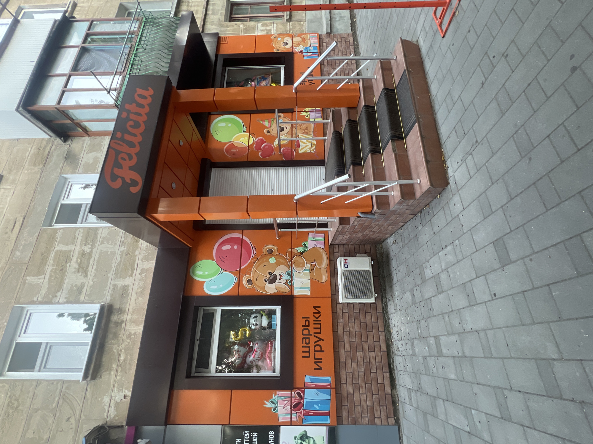 A storefront named "Felicita" is pictured in Bender, Moldova. The facade is painted in bright orange with brown trim. The store's name is in red cursive lettering above the entrance. The entrance features an orange canopy and steps with metal railings. The windows display balloons and cartoon bears. A sign with Russian text, "Шары Игрушки," translates to "Balloons Toys." An air conditioning unit is mounted on the brick wall below the window. The pavement in front of the store is made of gray interlocking stones.