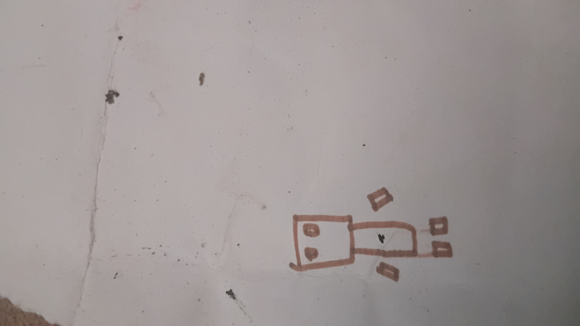 A crude, brown marker drawing of a stick figure is depicted on a white surface. The figure consists of a rectangular body with two circles inside representing eyes. Two rectangles extend from the left side of the body, suggesting arms or legs, and another two rectangles are positioned below the body. The drawing is slightly faded and smudged. The white surface appears to be paper, with some dirt specks and a faint crease running vertically through the right side of the image. The overall impression is of a child's simple drawing. The location of this image in Prabumulih, Indonesia, is not determinable from the visual information provided.