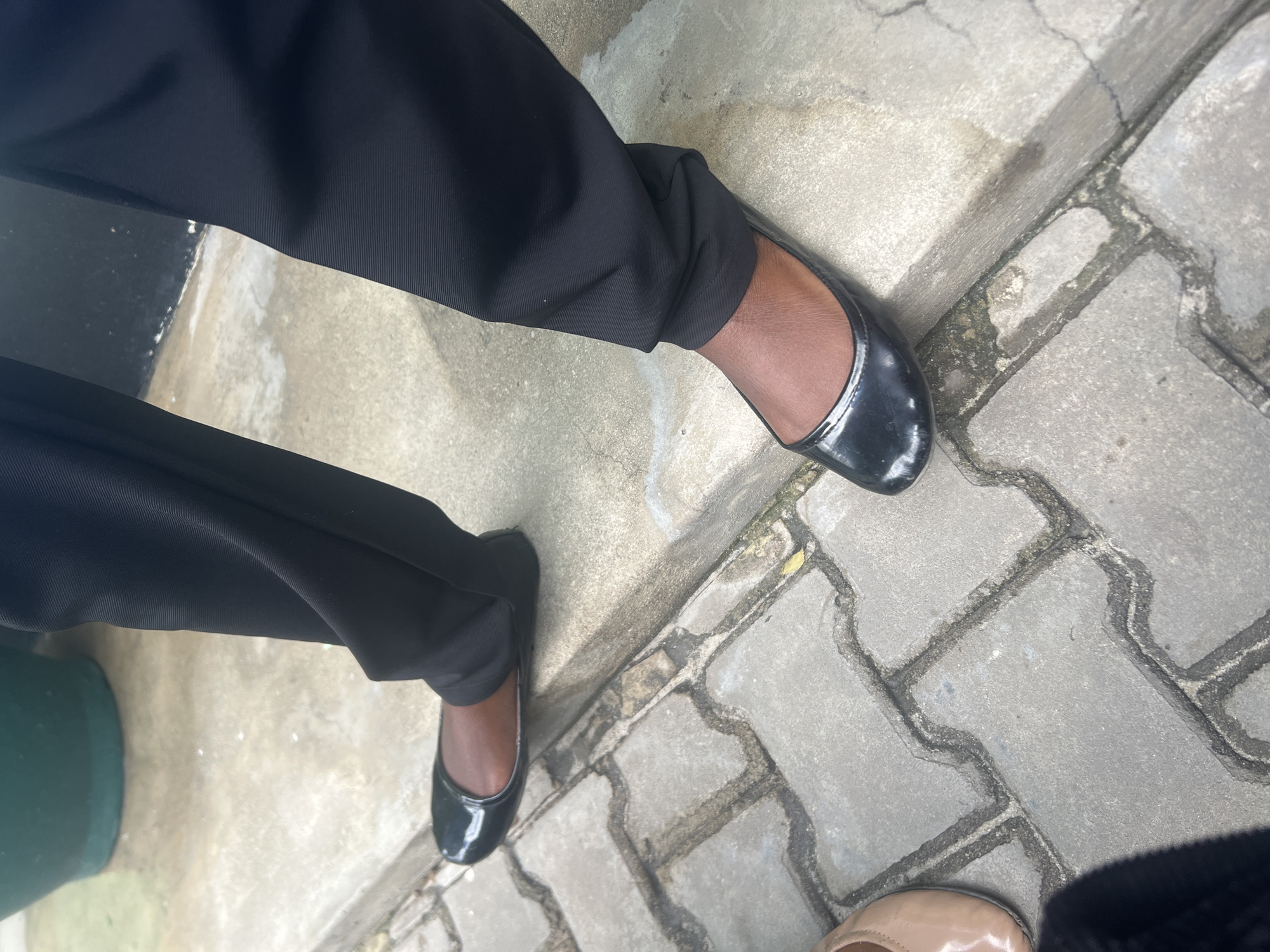 The image presents a low-angle, overhead view of the lower legs and feet of at least two individuals standing on an outdoor paved surface in what appears to be Dar es-Salaam, Tanzania. The setting suggests a pathway or sidewalk.
The main subject is a person wearing black trousers and black patent leather ballet flats. The trousers are a solid black, made from a smooth fabric that shows some creasing around the ankle. The shoes are shiny, reflecting light, with a classic rounded toe. The visible skin of the ankle indicates a medium to dark skin tone.
To the right and lower in the frame, the toe of a third shoe is visible, a light beige or tan ballet flat, also appearing to have a glossy finish. This suggests another person is standing nearby.
The ground surface is composed of two distinct materials. On the upper left, there is a smooth, lighter grey concrete slab. Transitioning from this, and covering most of the right side and bottom of the frame, are darker grey interlocking concrete pavers. These pavers have a unique "S" or wavy shape, creating an intricate pattern. The surfaces of both the concrete and pavers exhibit signs of wear, including visible cracks and some darker patches, possibly moss or dirt, within the paver joints. Several areas of the ground appear damp or wet.
In the lower-left corner, a dark green object, possibly a bucket or the base of a container, is partially visible. The lighting is bright and even, indicative of daytime. There is no visible text in the image. The scene is static, showing people standing in an everyday outdoor environment.