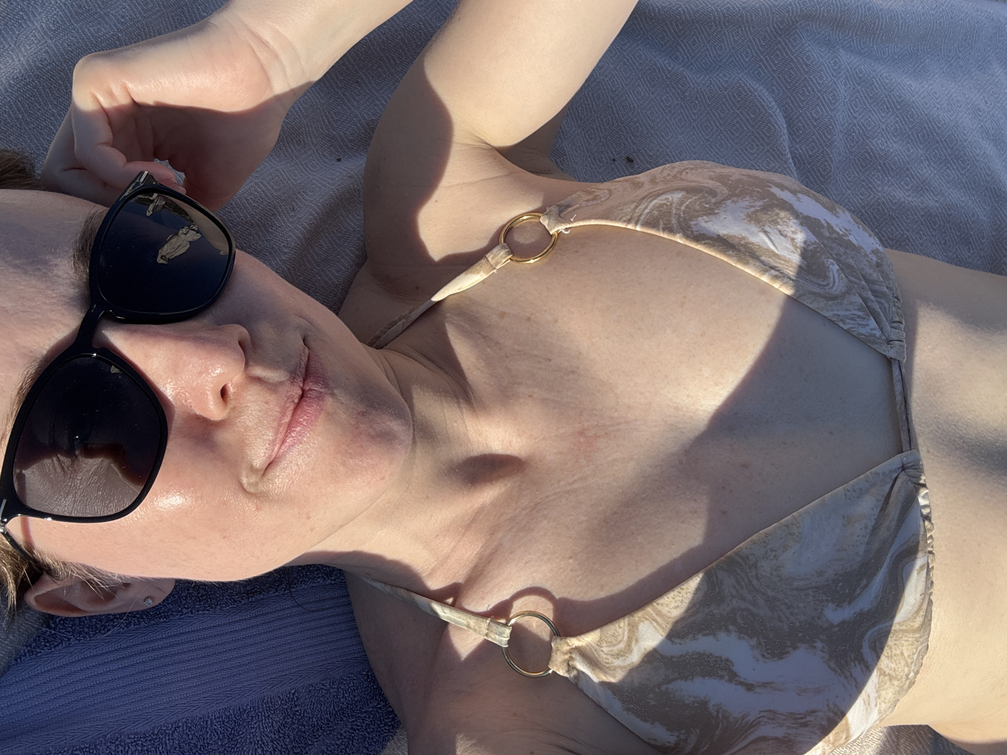 A person is lying on a towel outdoors. The person is wearing a bikini and sunglasses. The lighting suggests it is daytime. The image is a close-up and does not show the surrounding environment clearly.