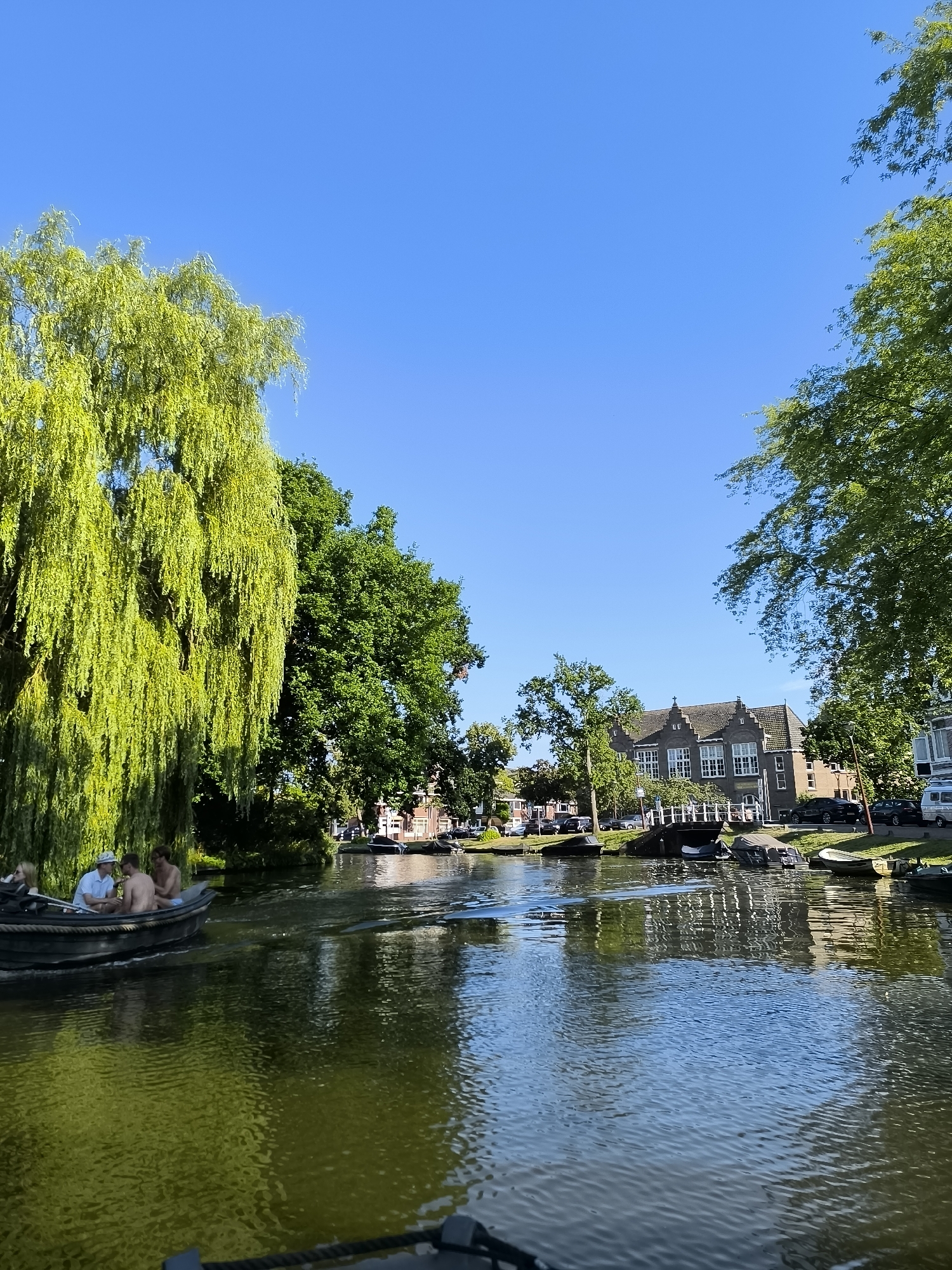 A photograph captures a canal scene in Alkmaar, Netherlands on a clear, sunny day. The water reflects the blue sky and the lush green foliage of trees lining the canal. In the foreground, a dark-colored boat navigates the waterway, carrying three individuals. Two men are seated in the boat, with one wearing a light-colored shirt and the other bare-chested. Another person, partially visible, is also in the boat. On the far bank of the canal, a street runs parallel to the water, lined with parked cars and residential buildings with pitched roofs. A white bridge spans the canal in the mid-ground. The overall impression is one of a tranquil summer day in a picturesque Dutch canal city.
