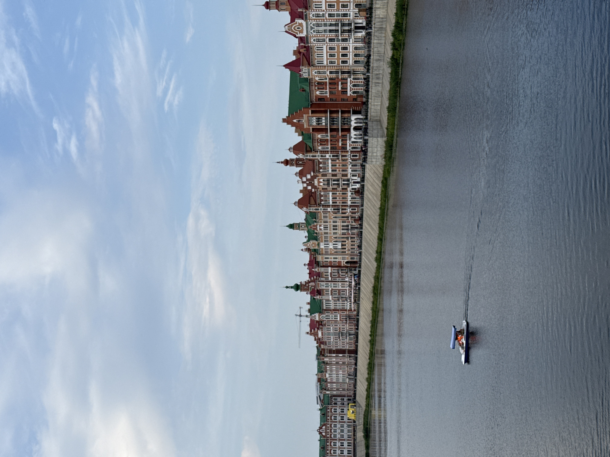This is a medium-shot, eye-level, outdoor, full shot photograph taken on a slightly overcast day. The scene is dominated by a body of water in the foreground, a long line of colorful, Dutch-style buildings in the middle ground, and a sky with scattered clouds in the background.
In the foreground, the water is a deep, dark blue with slight ripples on the surface. A small, white pedal boat with a blue canopy is visible in the left portion of the frame, moving from left to right. Two figures, wearing orange life vests, are seated in the boat.
The middle ground features a picturesque row of buildings that line the bank of the river. These buildings exhibit a variety of architectural styles, characterized by their brightly colored facades, often featuring red brick and white trim, and numerous windows. Many of the buildings have steeply pitched, green or red roofs, and some feature decorative spires or turrets. The buildings appear to be shops, restaurants, or residences, with awnings and outdoor seating visible in front of some establishments. A concrete embankment with a grassy edge separates the water from the buildings. A construction crane is visible in the distance behind the buildings.
The background consists of a pale blue sky with wispy white clouds, suggesting a temperate climate. The overall impression is one of a vibrant and charming European-inspired waterfront in Yoshkar-Ola, Russia.