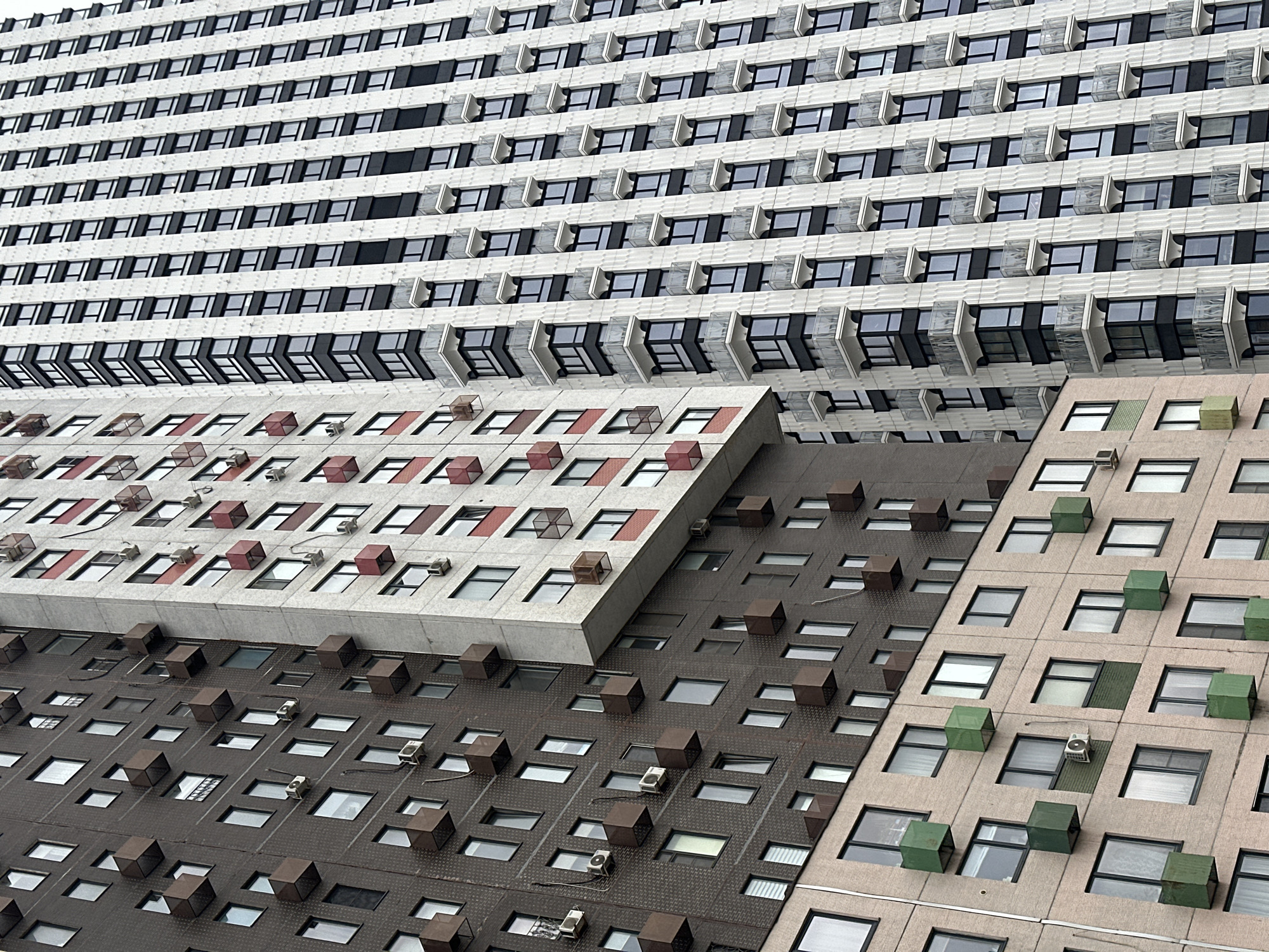 The image depicts the exterior of several connected, multi-story buildings in Khimki, Russia. The buildings have repeating patterns of windows and external fixtures. The uppermost building has a white facade with dark-framed windows, many of which feature external protruding fixtures. Beneath this building is a gray structure with red and white fixtures protruding from the facade near the windows. The lowest two structures are brown and beige, respectively, and feature window patterns with brown and green protruding fixtures. Several external air conditioning units are also visible on the building facades. There are no people visible in the image.