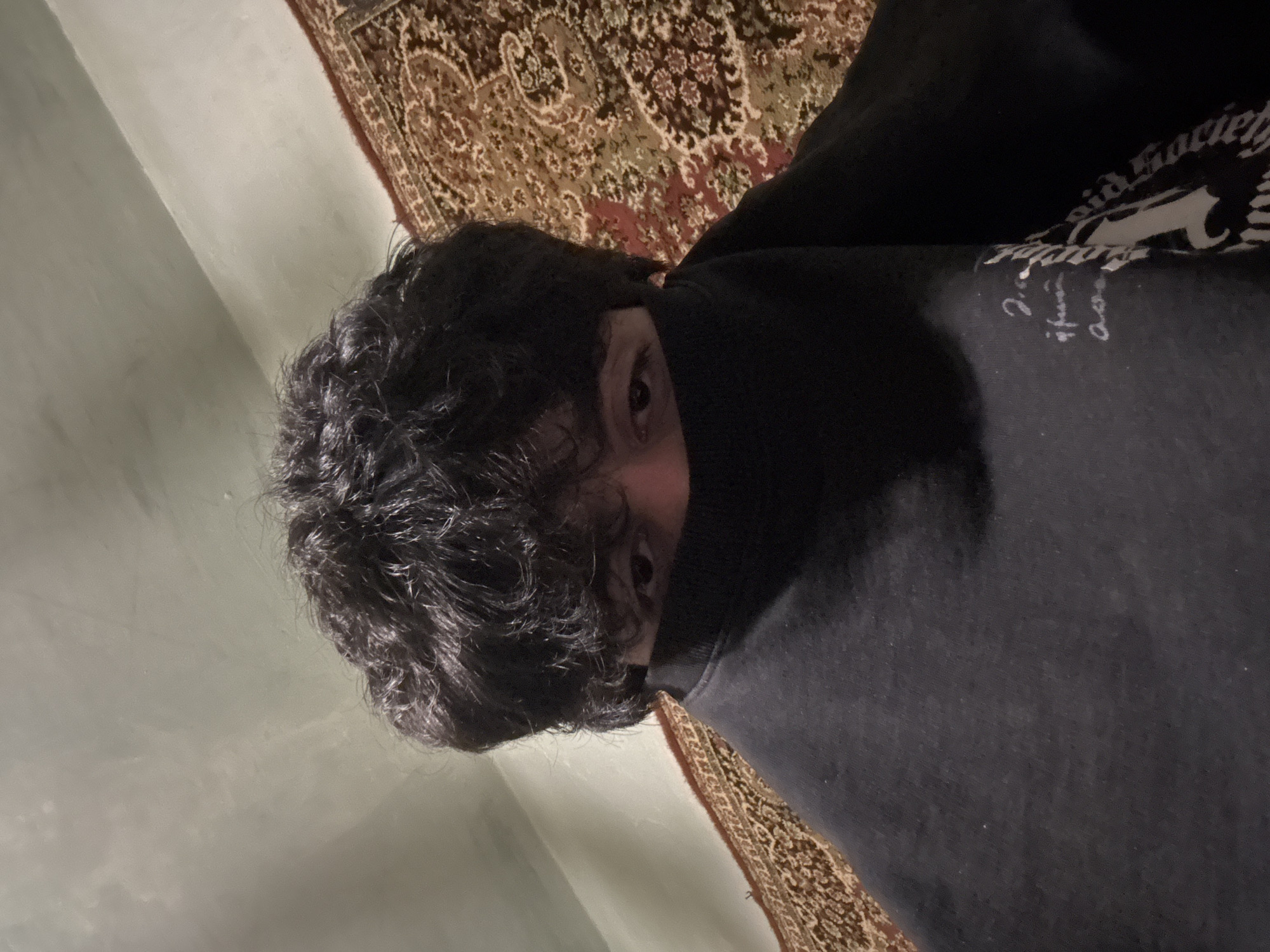 A person with dark curly hair is wearing a black hooded sweatshirt with writing on it. Only the eyes are visible. They are lying on a rug with a patterned design. The background suggests an indoor setting. No information indicates the location is Yerevan, Armenia.