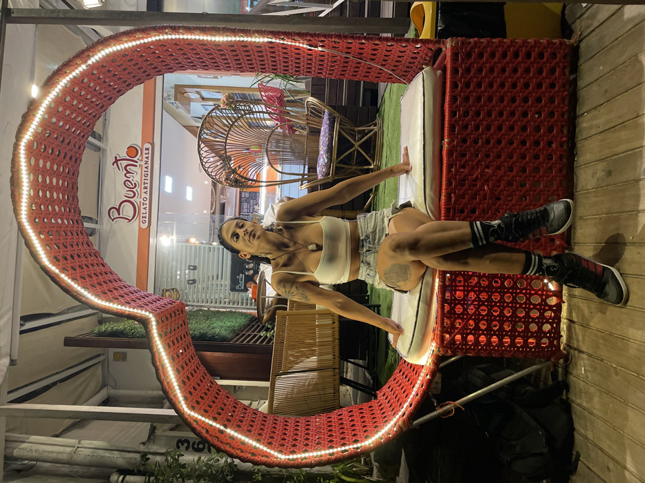 The image displays a person lounging in a unique red, illuminated seating structure, with a shopfront visible in the background. The photo is oriented vertically, though the original file seems to be rotated 90 degrees clockwise.
In the foreground, a person, likely female, is seated comfortably within a large, C-shaped lounge chair. This chair is made of a red, woven material, possibly rattan or wicker, and features a strip of white LED lights running along its inner edge, illuminating the structure. The person is leaning back against a white cushion placed inside the lounge. She has dark hair pulled back and is wearing a white spaghetti strap crop top, light-colored denim shorts, black high-top sneakers with white soles, and black socks with white patterns. She also wears a necklace with a pendant and has visible tattoos on her left arm and upper left thigh. Her left arm rests along the side of the red structure, while her right arm is bent. Her left leg is bent, and her right leg is extended forward, with both feet on the ground or a low platform. She is looking directly towards the camera with a direct gaze.
Behind the person, through a glass shopfront, the interior of a commercial establishment is visible. The shop's name, "Buono," is clearly displayed in a stylish orange script, with the words "Gelato Artigianale" underneath, indicating it is an artisan gelato shop. Inside the shop, several pieces of woven wicker or rattan furniture, including chairs and possibly a small table, can be seen, along with some green artificial turf used as decoration.
The ground or flooring in the immediate area around the red lounge chair is made of light-colored wooden slats. The presence of the illuminated LED lights on the structure and the brightly lit shop interior suggest that the photograph was taken during the evening or at night. The overall impression is that of a casual setting at a cafe or shop, with a unique and inviting seating area.