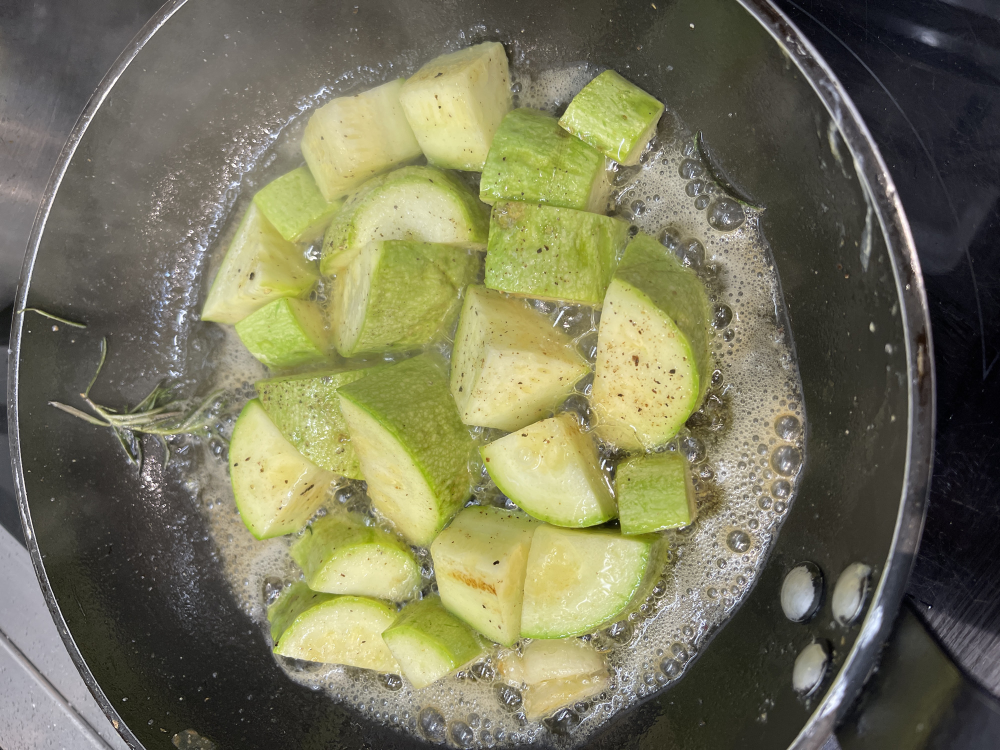 A close-up, overhead shot shows diced zucchini being sautéed in butter with a sprig of rosemary in a black frying pan. The butter is bubbling, and there are seasoning specks on the zucchini. The frying pan is on a black stovetop. The location of the image is not visible. There is no information indicating that the image was taken in Belgrade, Serbia.