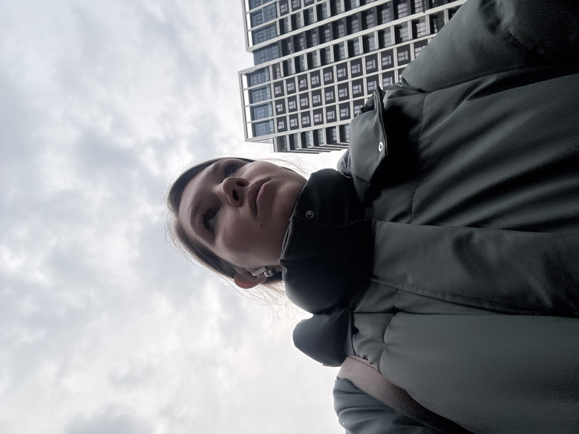 A woman in a dark green puffer jacket looks up at a tall, modern building with a grid-like facade of windows. The sky is overcast. The photo seems to be taken from a low angle. It could be in Belgrade, Serbia, due to the architecture and overall feel of the image, but confirmation would require additional visual cues specific to Belgrade.