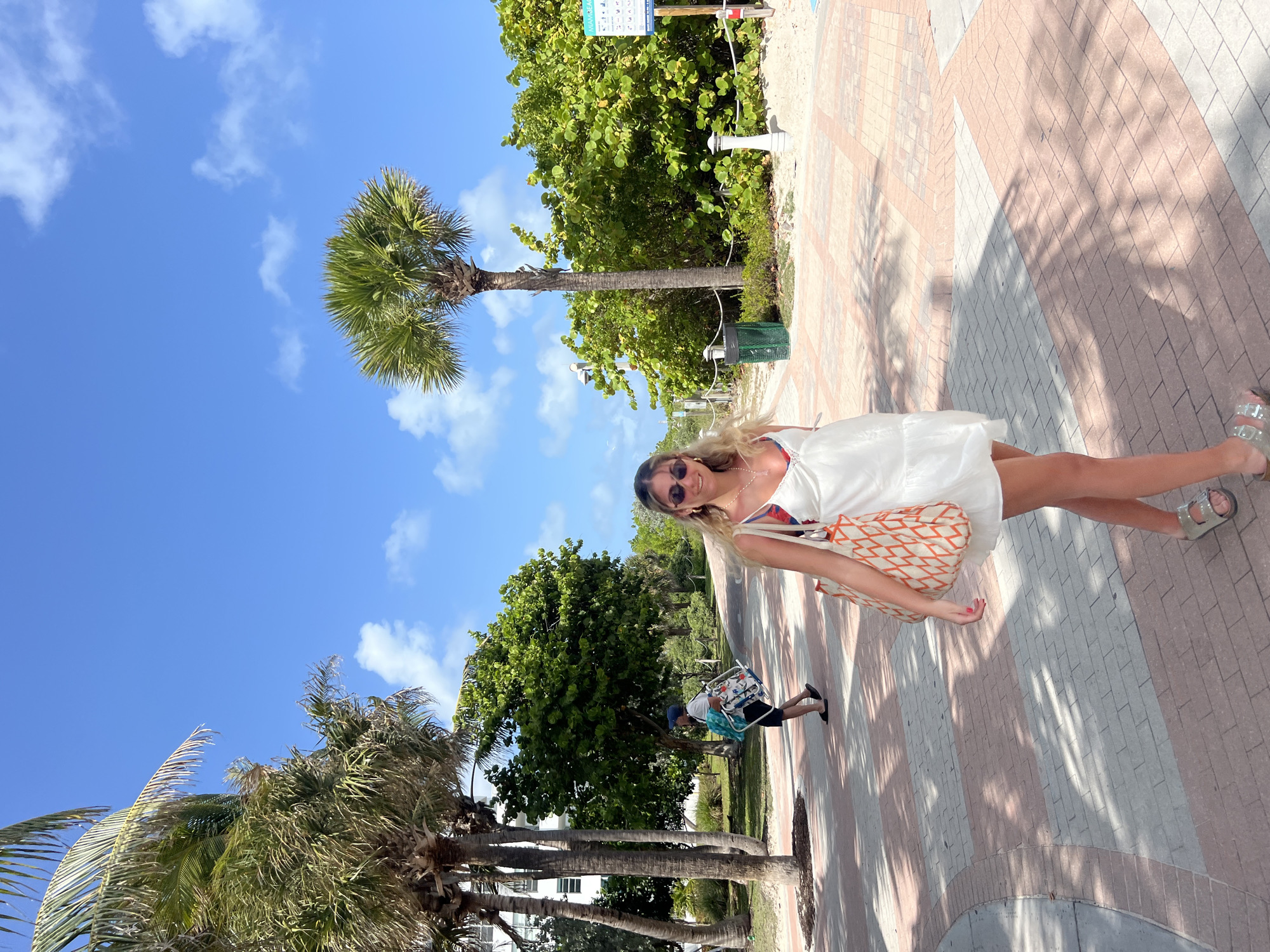A slightly tilted, medium shot shows a woman in a white dress, sunglasses, and a patterned bag on a brick pathway in Miami Beach, United States of America. The pathway is patterned with alternating rows of red and white bricks, with palm tree shadows cast on the bricks. Behind the woman, palm trees and lush green foliage are visible against a blue sky dotted with clouds. A man carrying a beach chair is also visible.