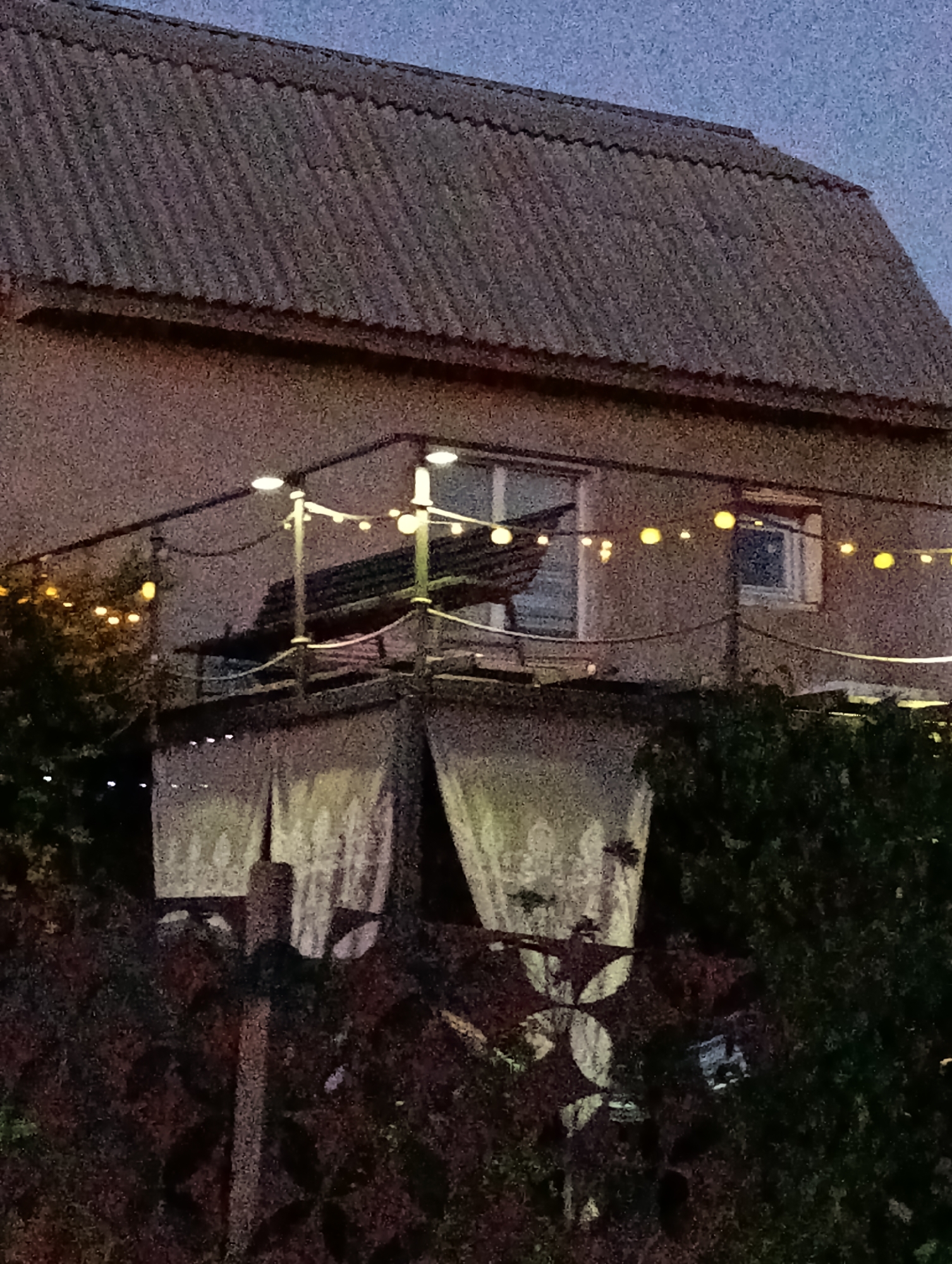 The image shows the exterior of a building with a corrugated roof. There is a balcony with a railing and string lights, and two large white curtains hanging below the balcony. The scene appears to be set in the city of Roghi, Moldova, as indicated by the provided context. The environment is dark, suggesting it is nighttime or dusk. There are no people visible in the image.