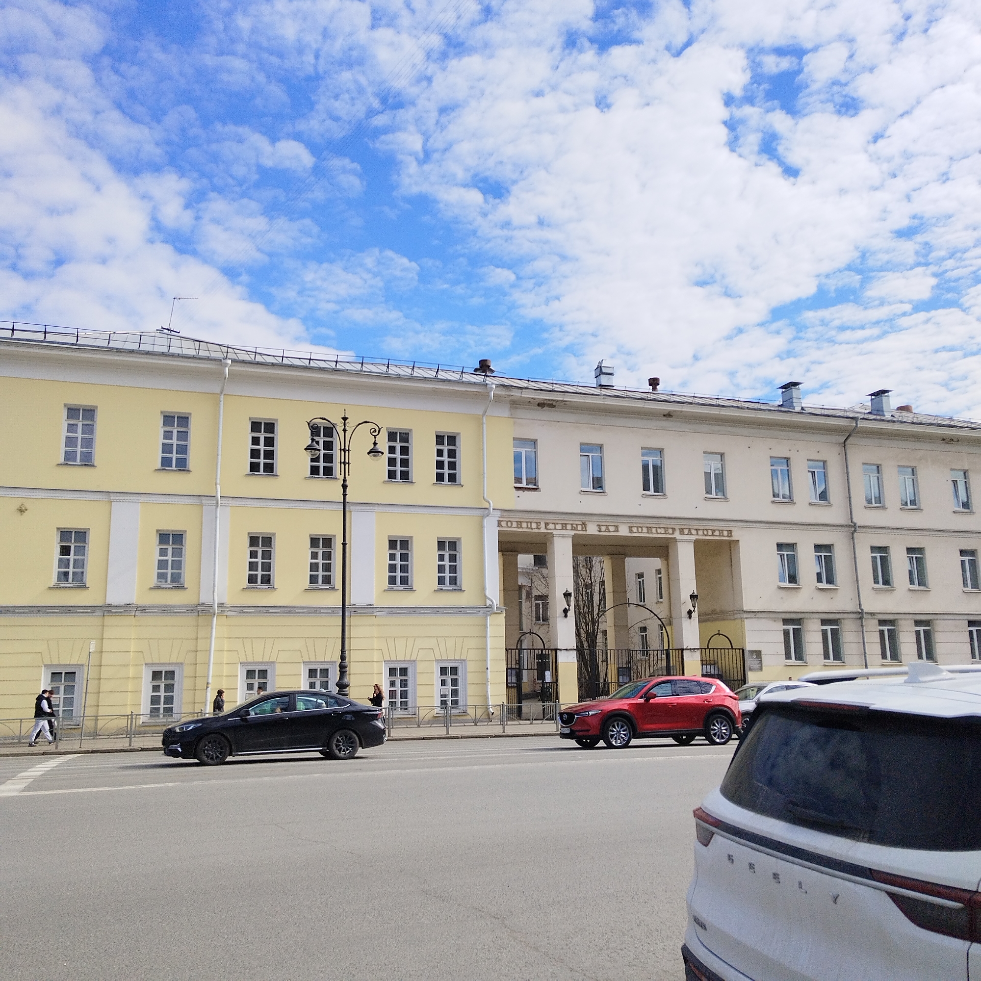 The image depicts a street scene in Yekaterinburg, Russia, featuring a building with two distinct architectural styles. The left portion of the building is yellow with multiple windows, while the right portion is a lighter color with a large arched entrance in the center, marked above the arch with the words "Концертный зал консерватории" (Concert Hall of the Conservatory).
Three cars are visible on the street: a black sedan traveling left, a red SUV moving right, and the rear of a white SUV in the foreground. There are a few pedestrians visible on the left of the frame. The sky is blue with scattered white clouds. A lamppost stands on the sidewalk next to the yellow building.