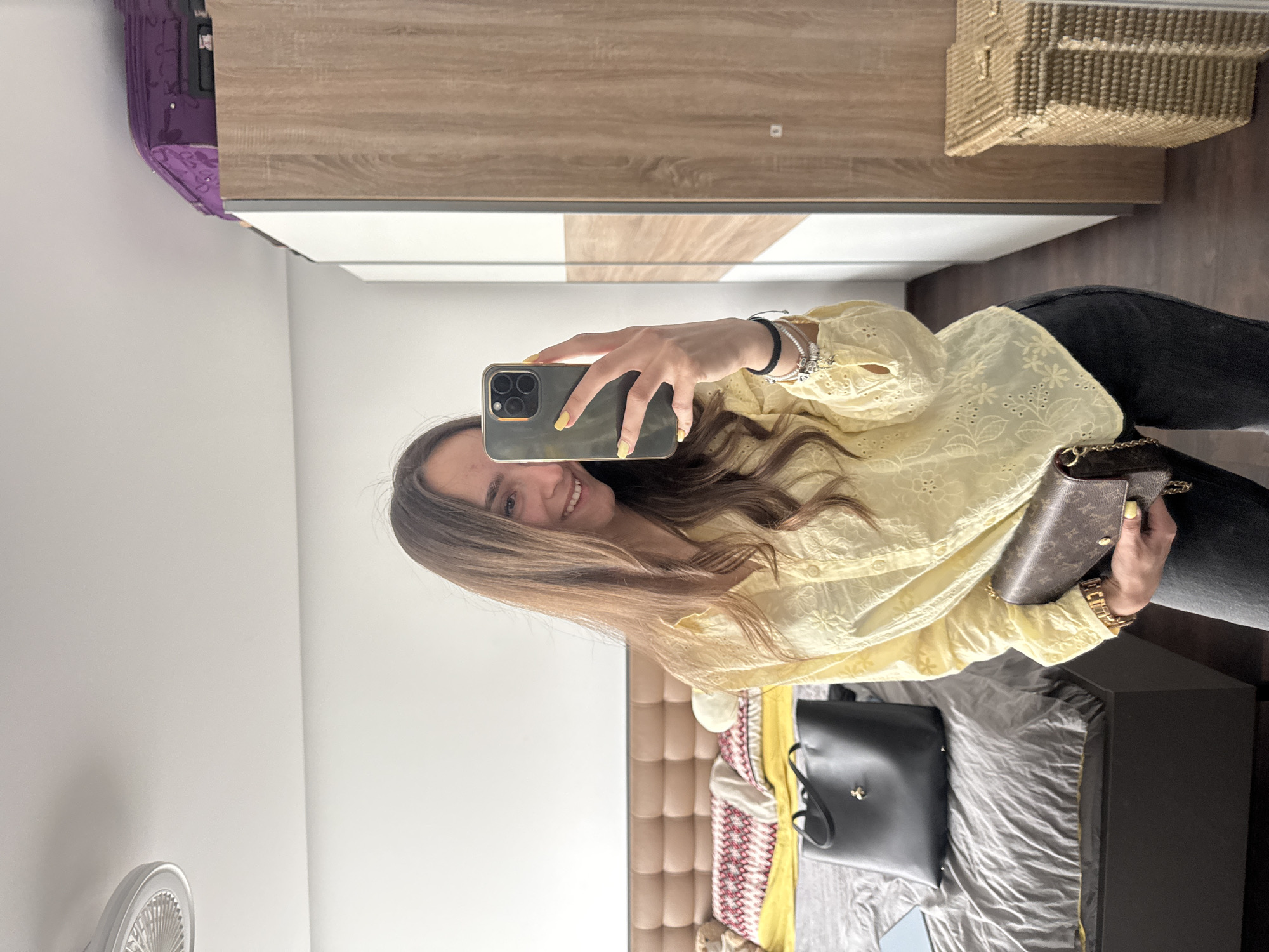 The image depicts a young woman taking a selfie in what appears to be a bedroom. The woman is fair-skinned with long brown hair and is wearing a yellow blouse with eyelet detailing and black pants. She is holding a smartphone in her left hand, with a brown and gold chain purse visible. The room contains a bed with a grey comforter, a black tote bag, a headboard, a luggage in the top left, and a dresser with a woven box on top. The setting indicates an interior space, likely in a residential area. The city of Sofia, Bulgaria cannot be determined from the image.