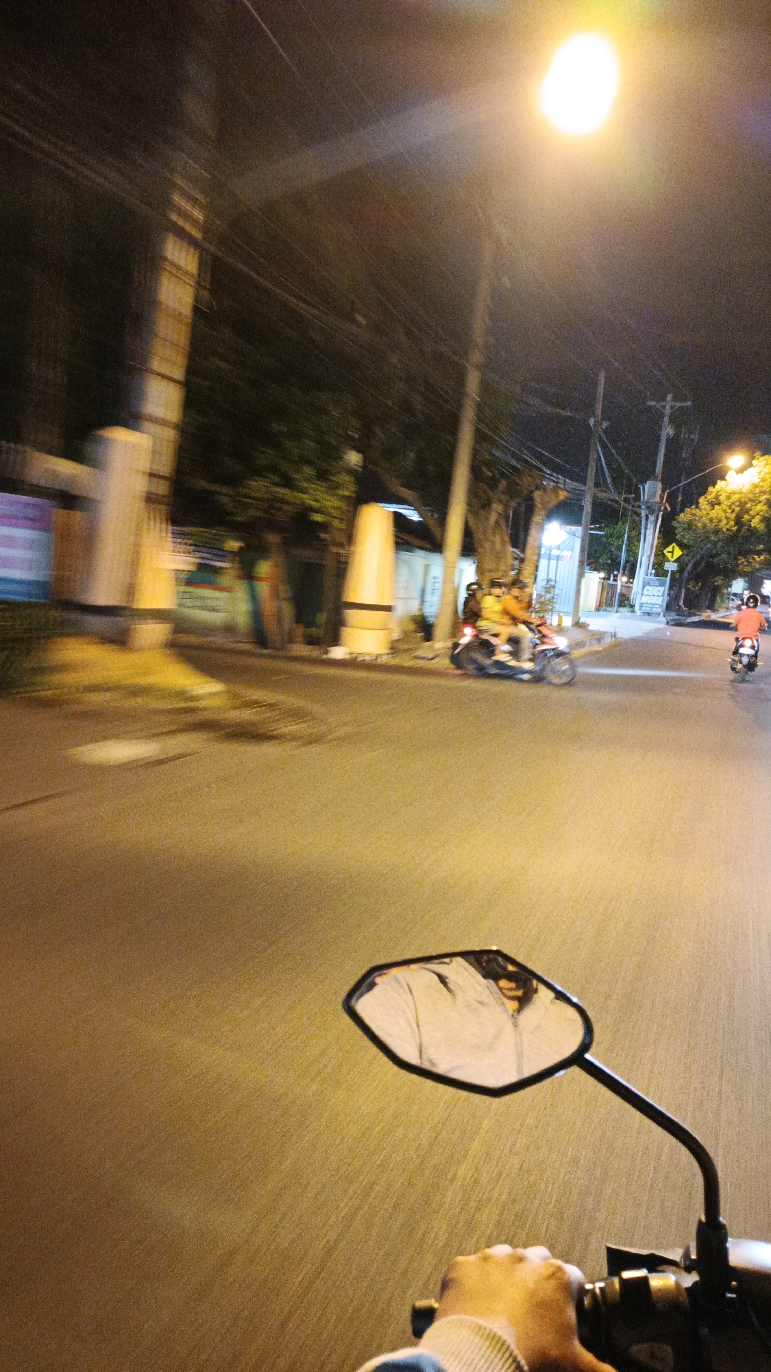 The image captures a nighttime street scene in Surakarta, Indonesia, with a prominent streetlamp illuminating the area. The perspective is from a motorcycle, with the rider's hand gripping the handlebar and a side mirror reflecting the rider's face and jacket. Several other motorcycles are visible, some with passengers, traveling on the asphalt road. The background shows buildings, trees, and utility poles, all lit by the streetlamp and other ambient light sources. The motion blur suggests the motorcycles are in motion, contributing to the dynamic feel of the image. The overall scene depicts a typical urban environment at night.